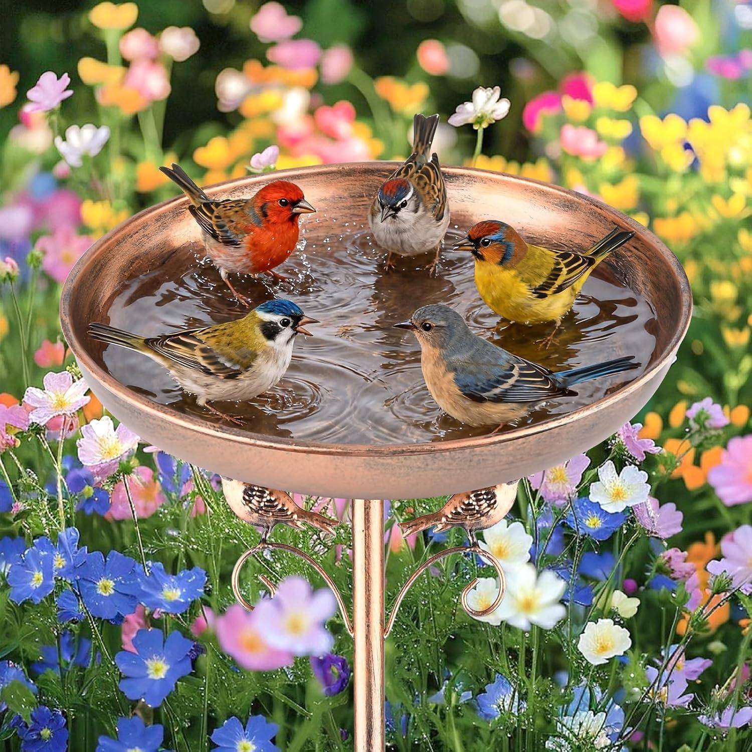 Fünf bunte Vögel sitzen in einem mit Wasser gefüllten Vogelbad, umgeben von blühenden rosa, weißen und blauen Blumen in einem Garten.