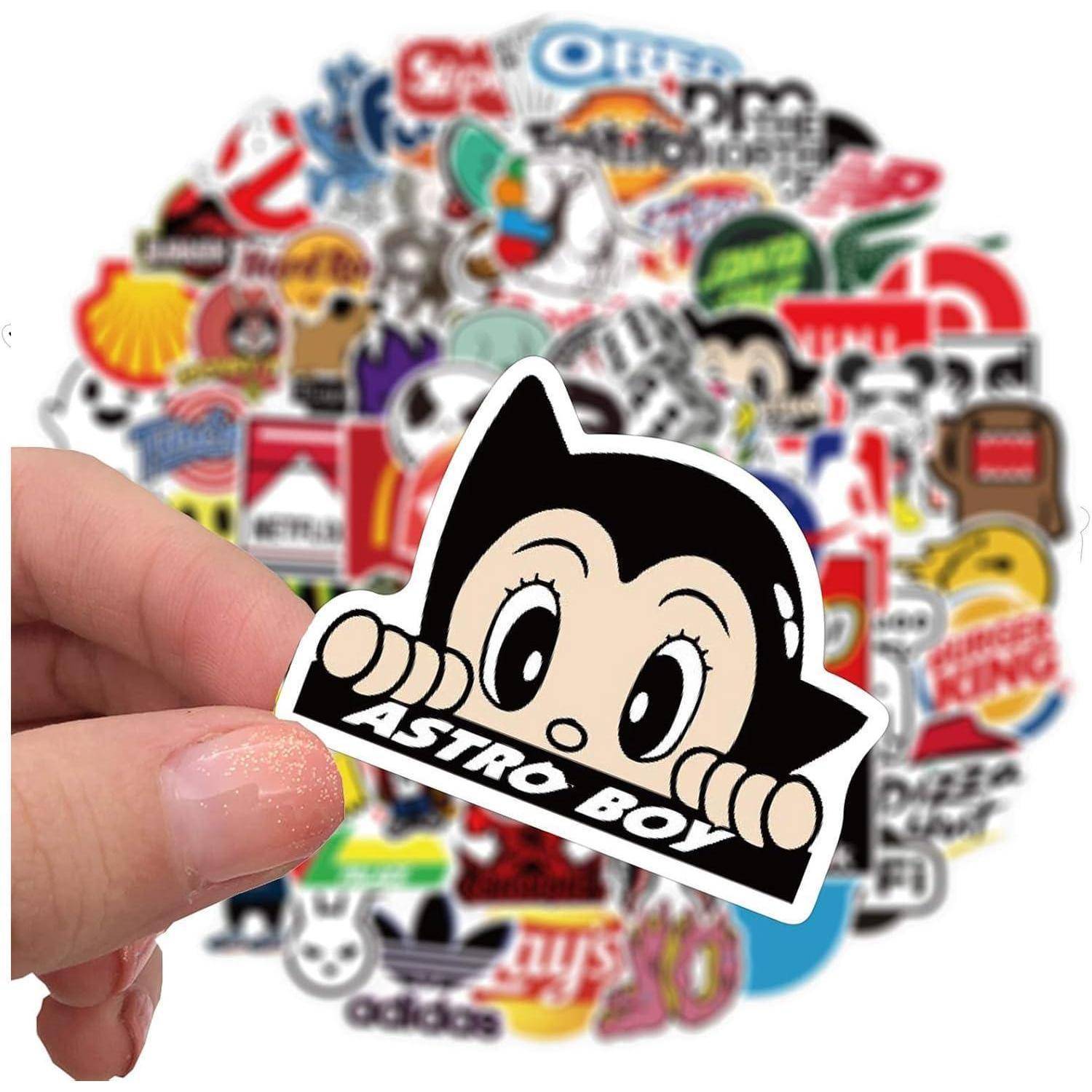 Hand, die einen "ASTRO BOY""-Aufkleber mit dem Gesicht der Cartoon-Figur hält; ein unscharfer Haufen assortierter Aufkleber füllt den Hintergrund.