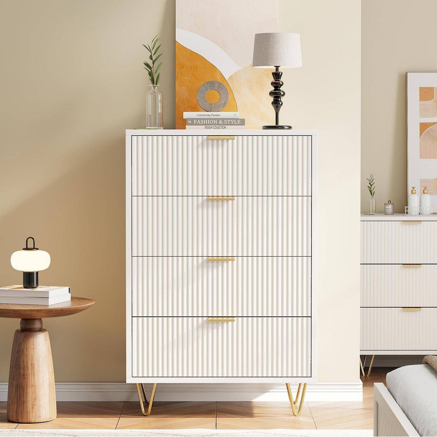 Modernes Schlafzimmerszenario mit einer weißen gerippten Kommode mit vier Schubladen auf goldenen Beinen, die mit Dekoration und einer Lampe bestückt ist, neben einer passenden Kommode und einem Beistelltisch.