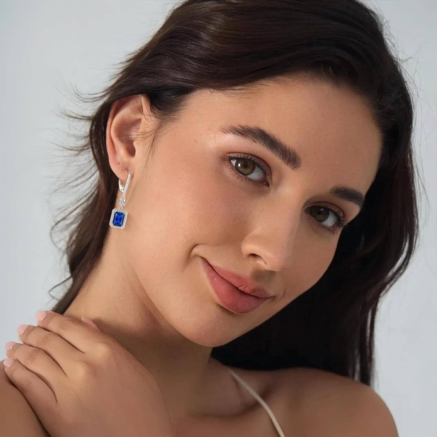 Porträt einer Frau mit sanftem Lächeln, die einen blauen Edelsteinohring trägt und ihre Schulter gegen einen schlichten, hellen Hintergrund berührt.