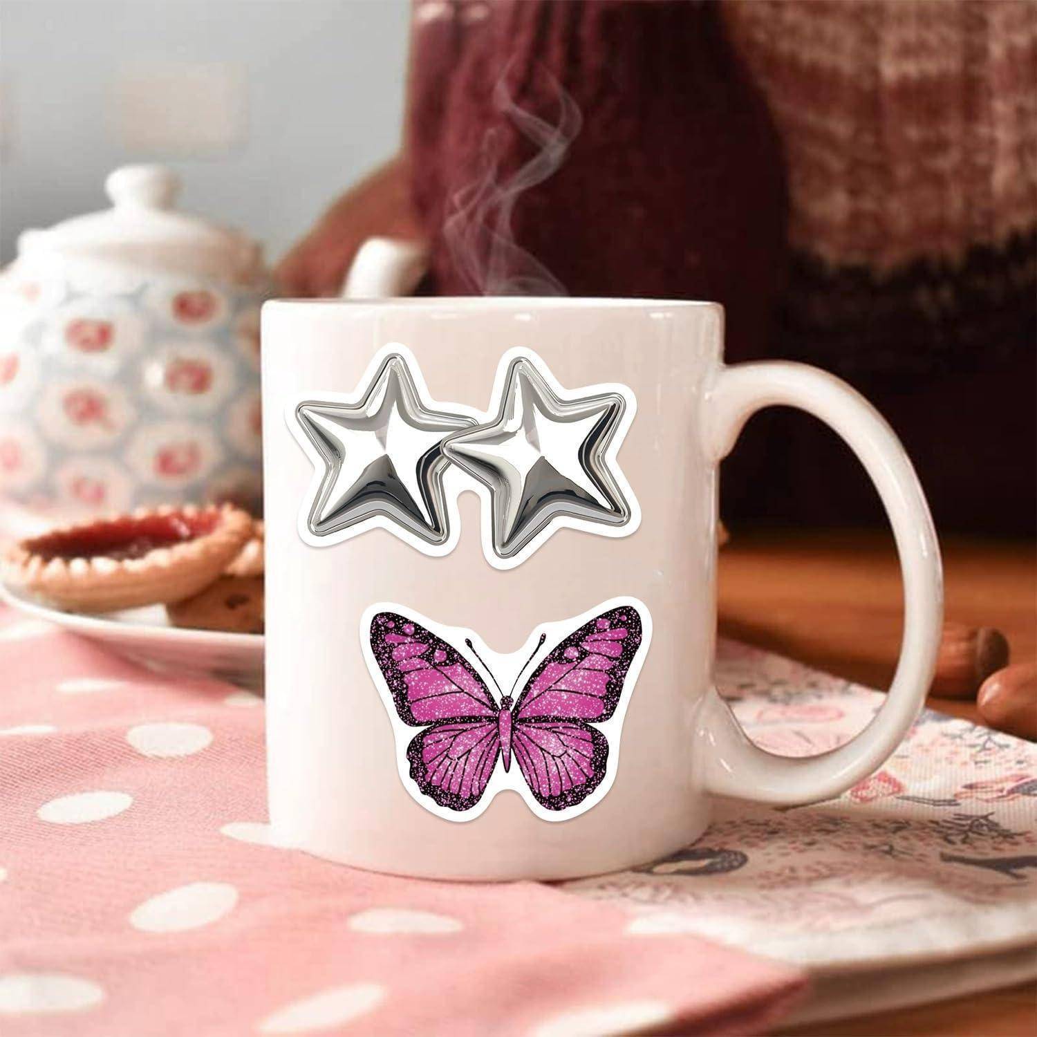 Weiße Tasse mit zwei silbernen Sternenaufklebern und einem rosa Schmetterlingsaufkleber, dampfend auf einem rosa Tisch mit weißen Punkten, mit einer Teekanne und einem Gebäck im Hintergrund.