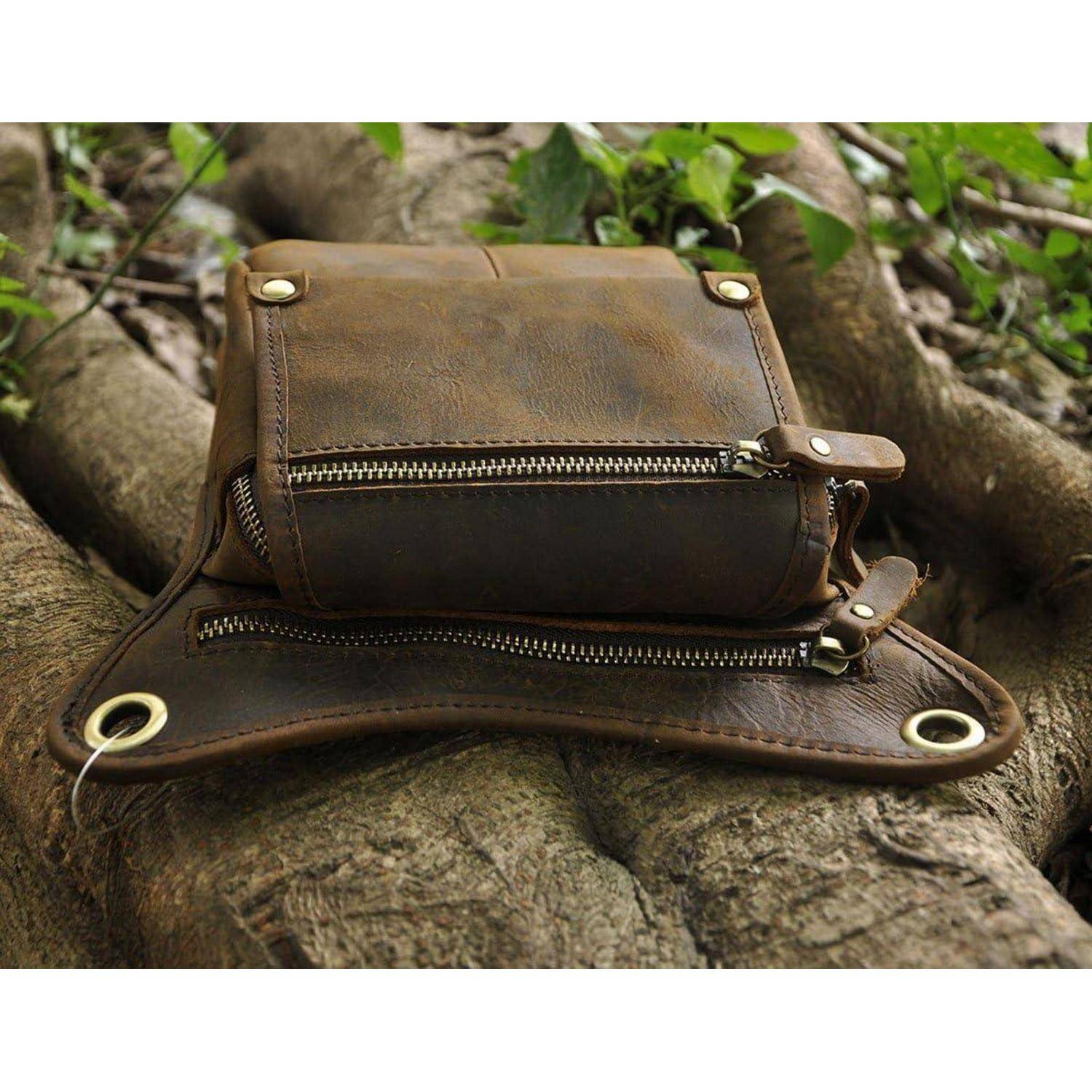 Braunes Lederetui mit Reißverschluss oder kleine Tasche, das/die auf Baumwurzeln im Freien ruht, mit Messingösen und Metallbeschlägen.