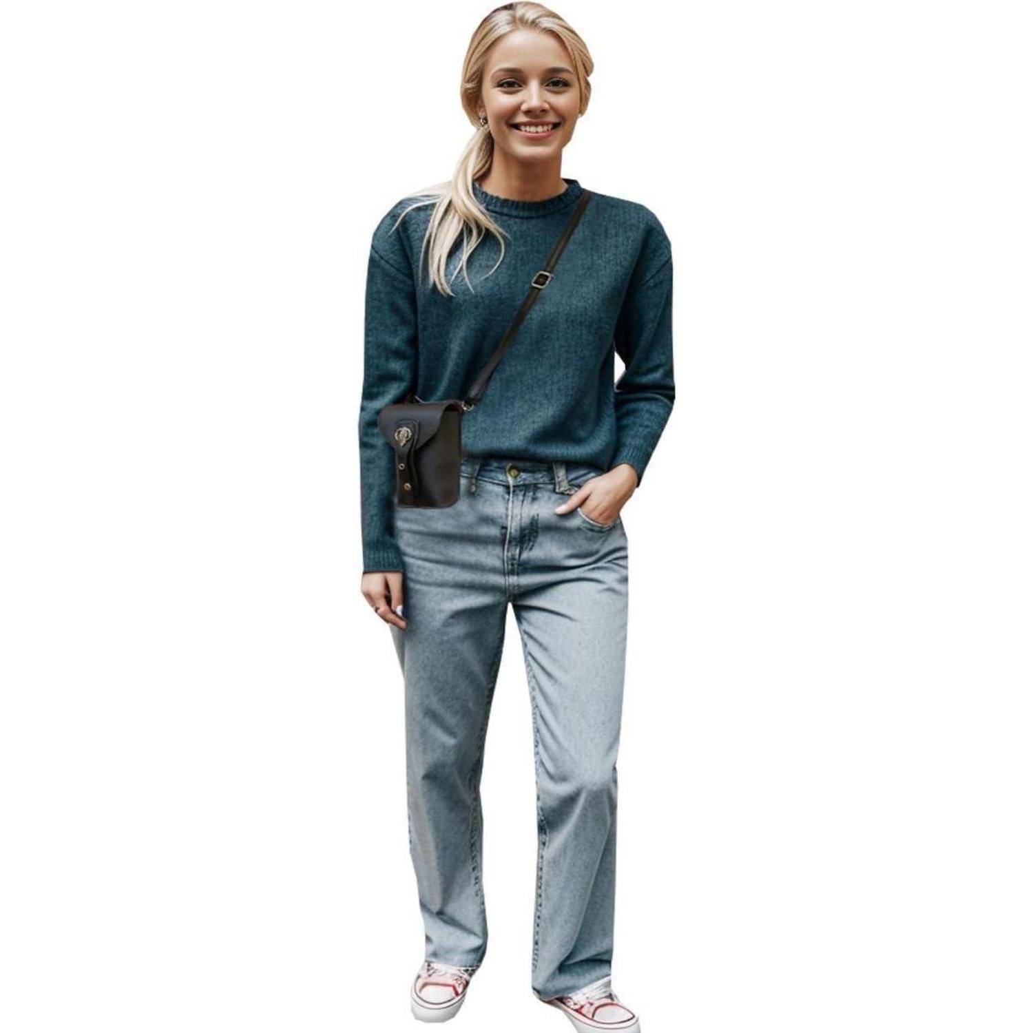 Eine lächelnde Frau, die in voller Länge vor weißem Hintergrund steht, trägt einen blaugrünen Pullover, helle Jeans, Sneaker und eine kleine Umhängetasche.