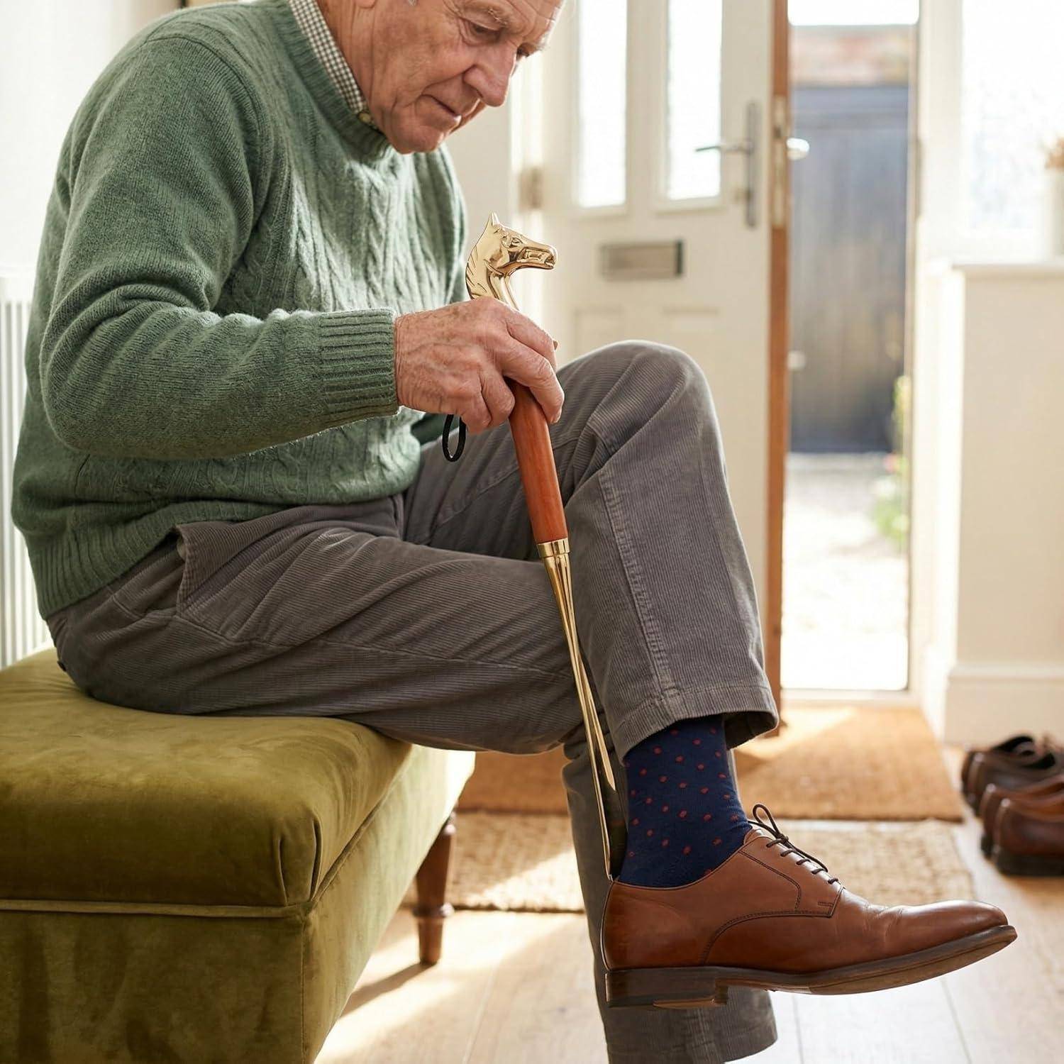 Ein älterer Mann sitzt auf einer grünen Bank und benutzt einen Schuhanzieher, um einen braunen Anzugschuh in einem hellen Eingangsbereich anzuziehen.
