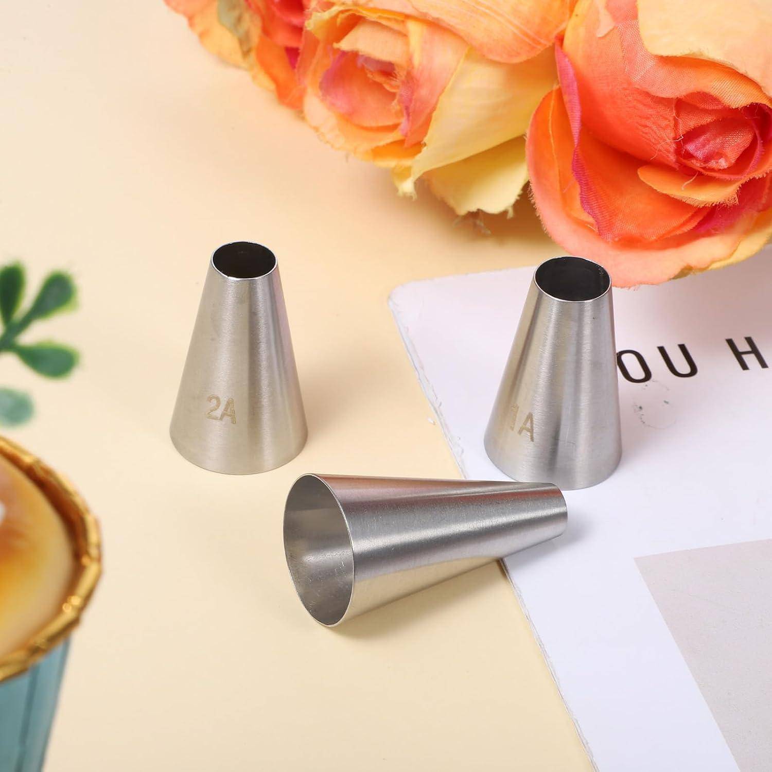 Drei Metallspritzbeutel für Tortendekoration, angeordnet auf einer Pastelloberfläche neben Blumen und einem teilweise sichtbaren Buch.