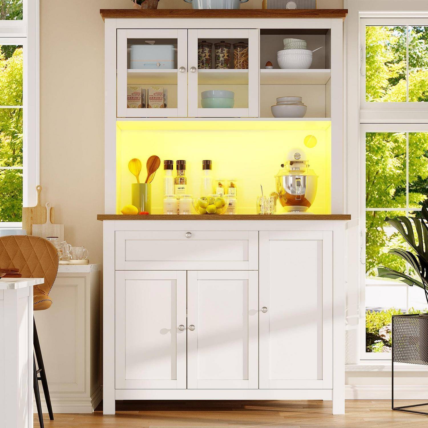 Weiße Küchenvitrine mit Glasschiebetüren, beleuchtete gelbe Rückwand, Arbeitsplatte mit Gläsern und Mixer, neben hellen Fenstern in einem sonnigen Zimmer