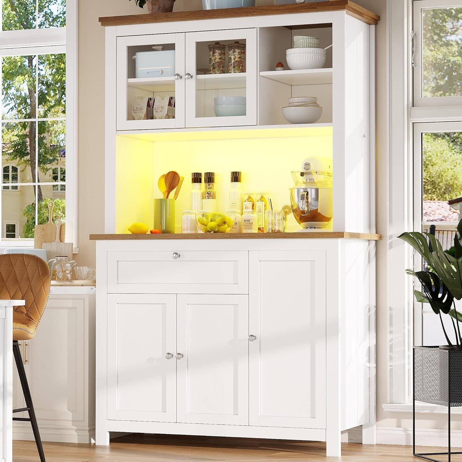 Weißes Küchenbuffet mit Glastüren und gelb beleuchteten offenen Regalen, die Schüsseln, Gläser, Küchenutensilien, Zitronen und eine Küchenmaschine neben sonnigen Fenstern ausstellen.