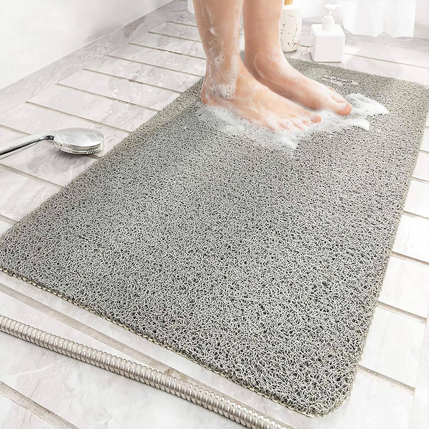 Person wäscht sich die Füße, während sie auf einer grauen Badematte steht, mit Seifenschaum und einem Duschkopf in der Nähe auf einem gekachelten Badezimmerboden.