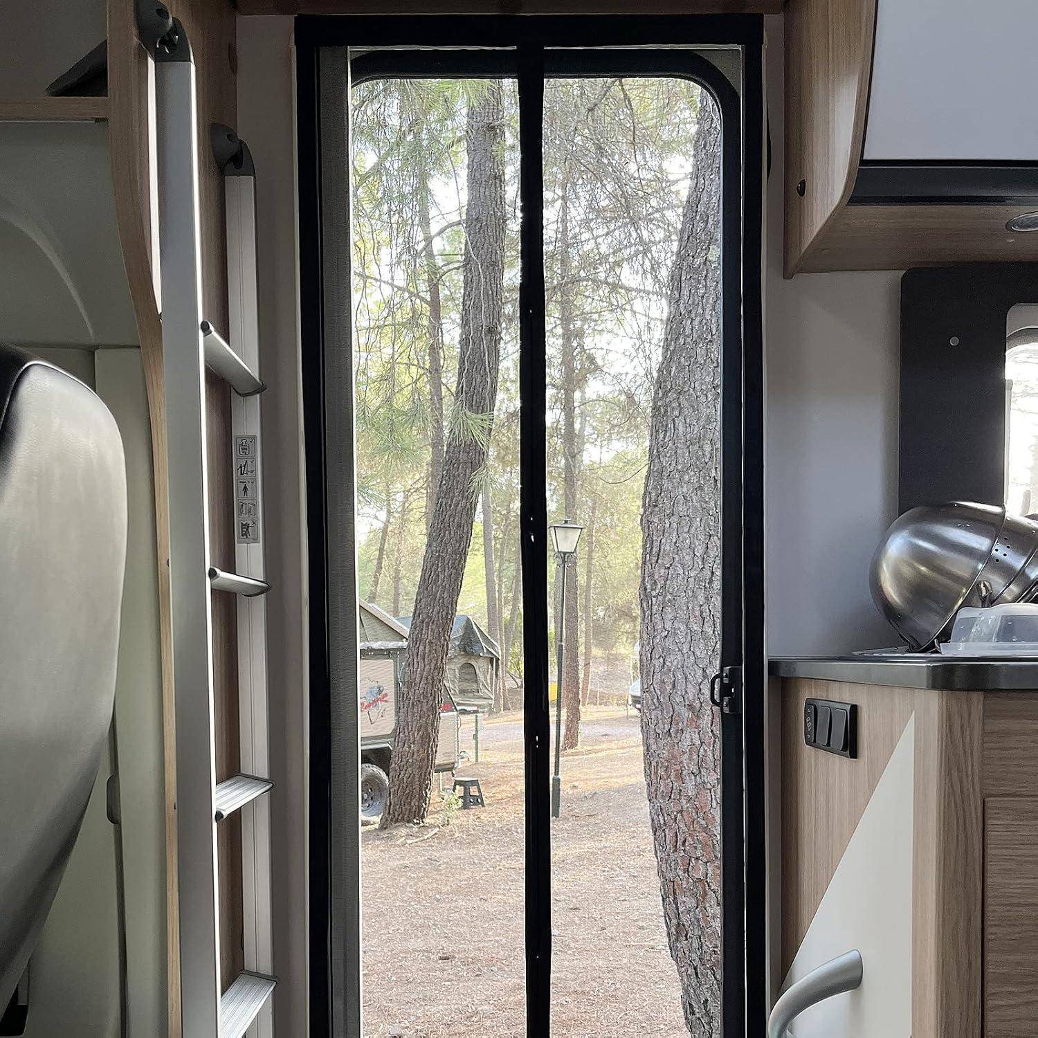 Blick von innen durch eine Wohnmobiltür durch eine Fliegengittertür auf einen bewaldeten Campingplatz mit Bäumen, einer Lampe und einem kleinen Unterstand außen.