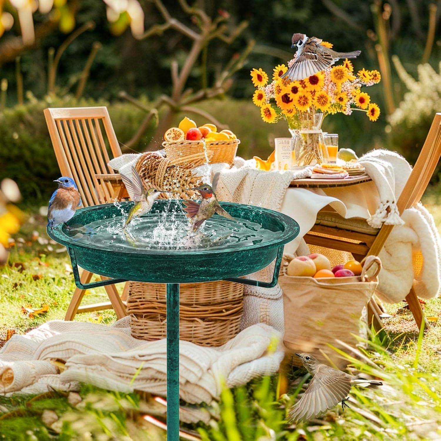 Vögel planschen in einem Gartenvogelbad neben einem Picknicktisch, der auf einem sonnigen Rasen mit Obst, Blumen und Korbkörben eingedeckt ist.