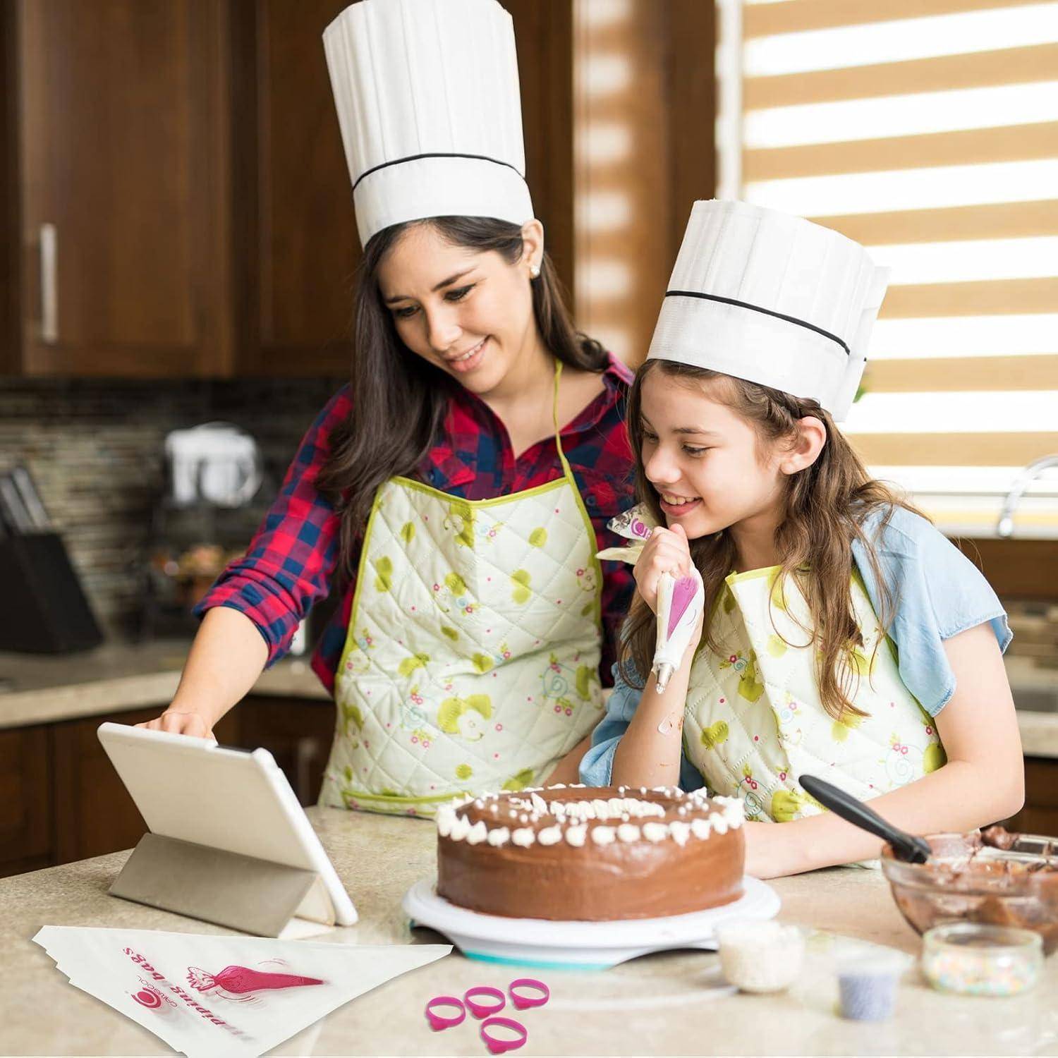 Mutter und Tochter in Kochmützen und Schürzen dekorieren einen Schokoladenkuchen, während sie einem Rezept auf einem Tablet in einer Küche folgen.