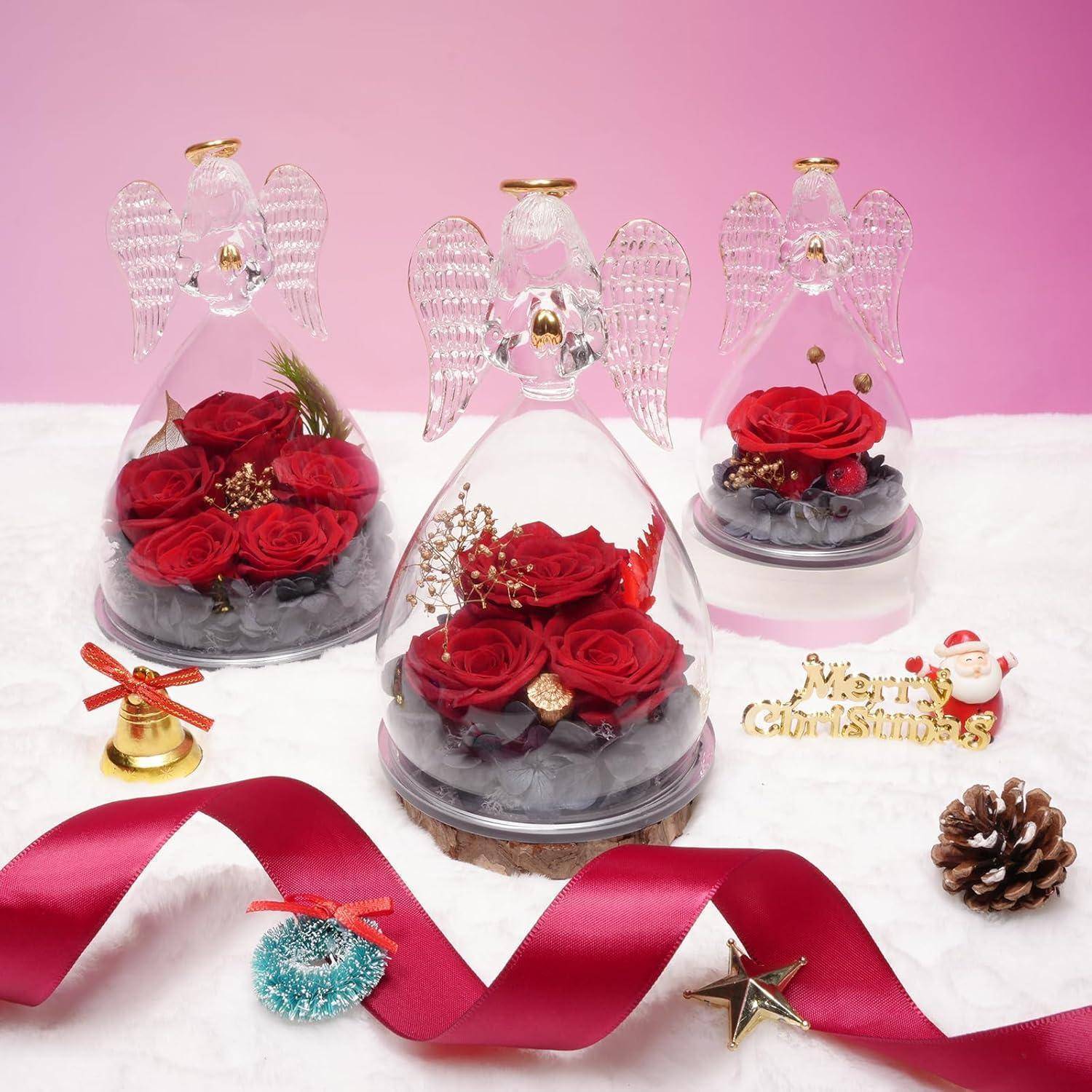 Drei Glasengelfiguren mit roten Rosen und dunklen Steinen, umgeben von Weihnachtsdekoration, einschließlich Schleife, Glocke, Tannenzapfen und 
