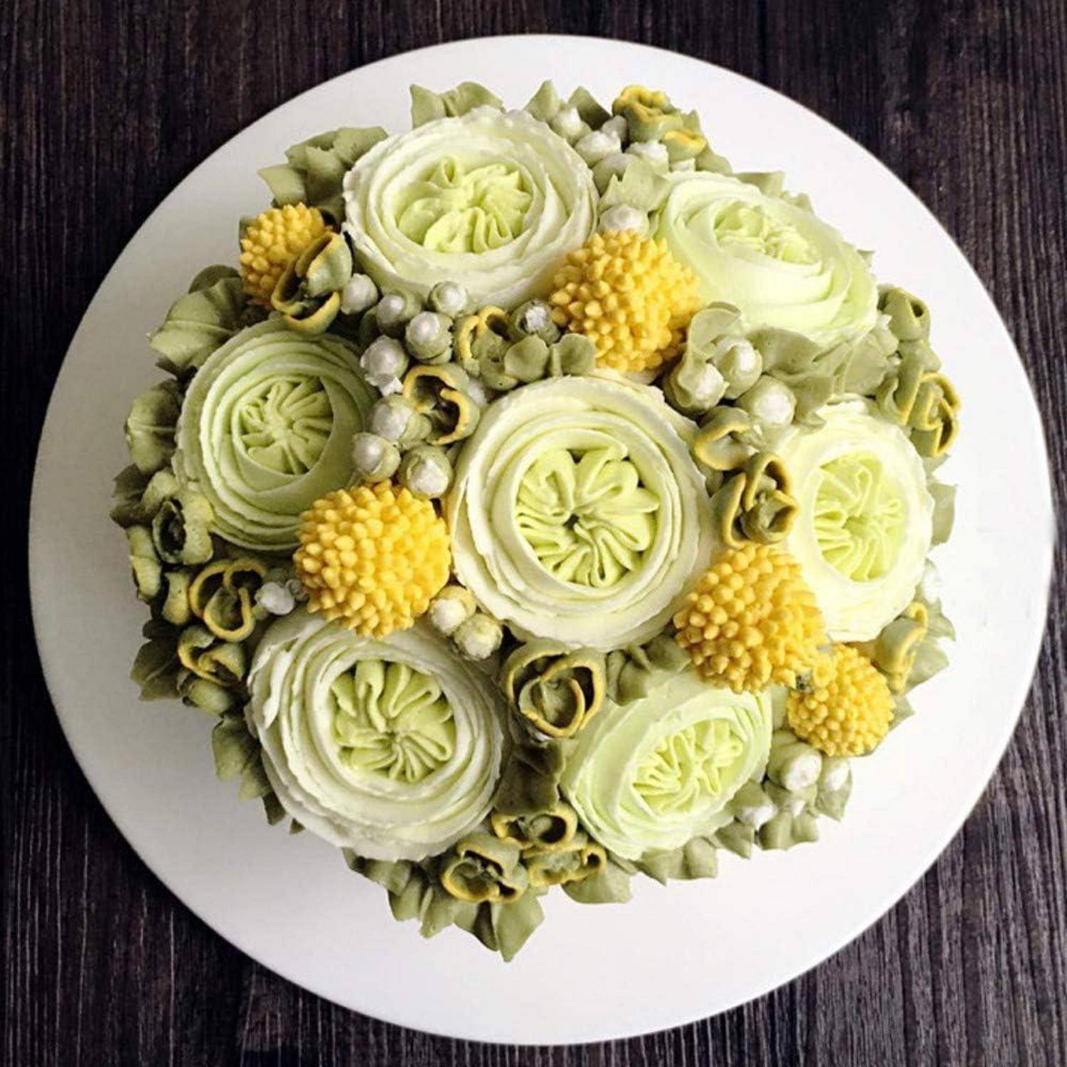 Draufsicht eines runden Kuchens, der mit hellgrünen und gelben Buttercreme-Blüten auf einem weißen Teller verziert ist.
