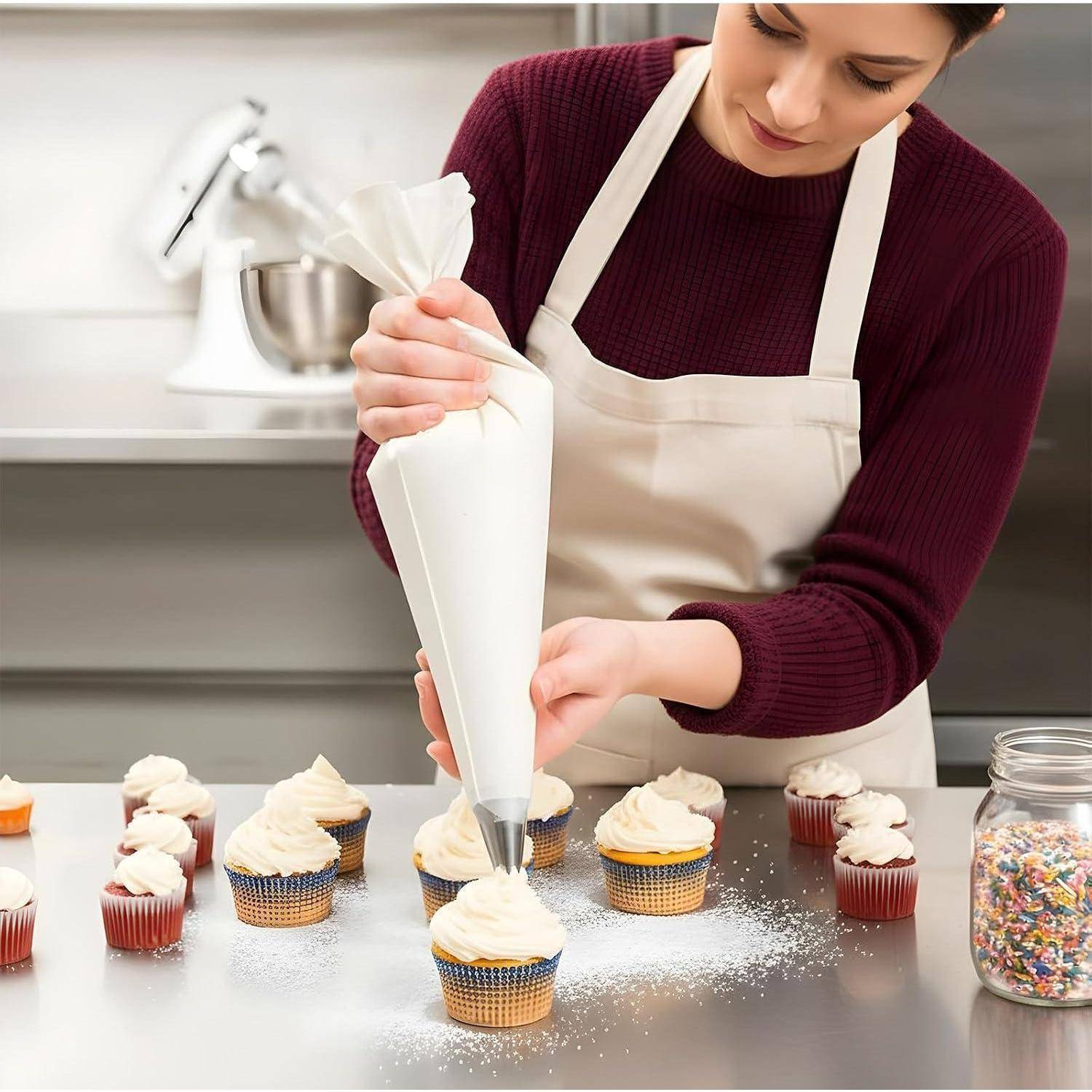 Frau in Schürze, die Zuckerguss auf Cupcakes auf einer Arbeitsplatte spritzt, mit mehreren dekorierten Cupcakes und einem Glas mit Streuseln in der Nähe.