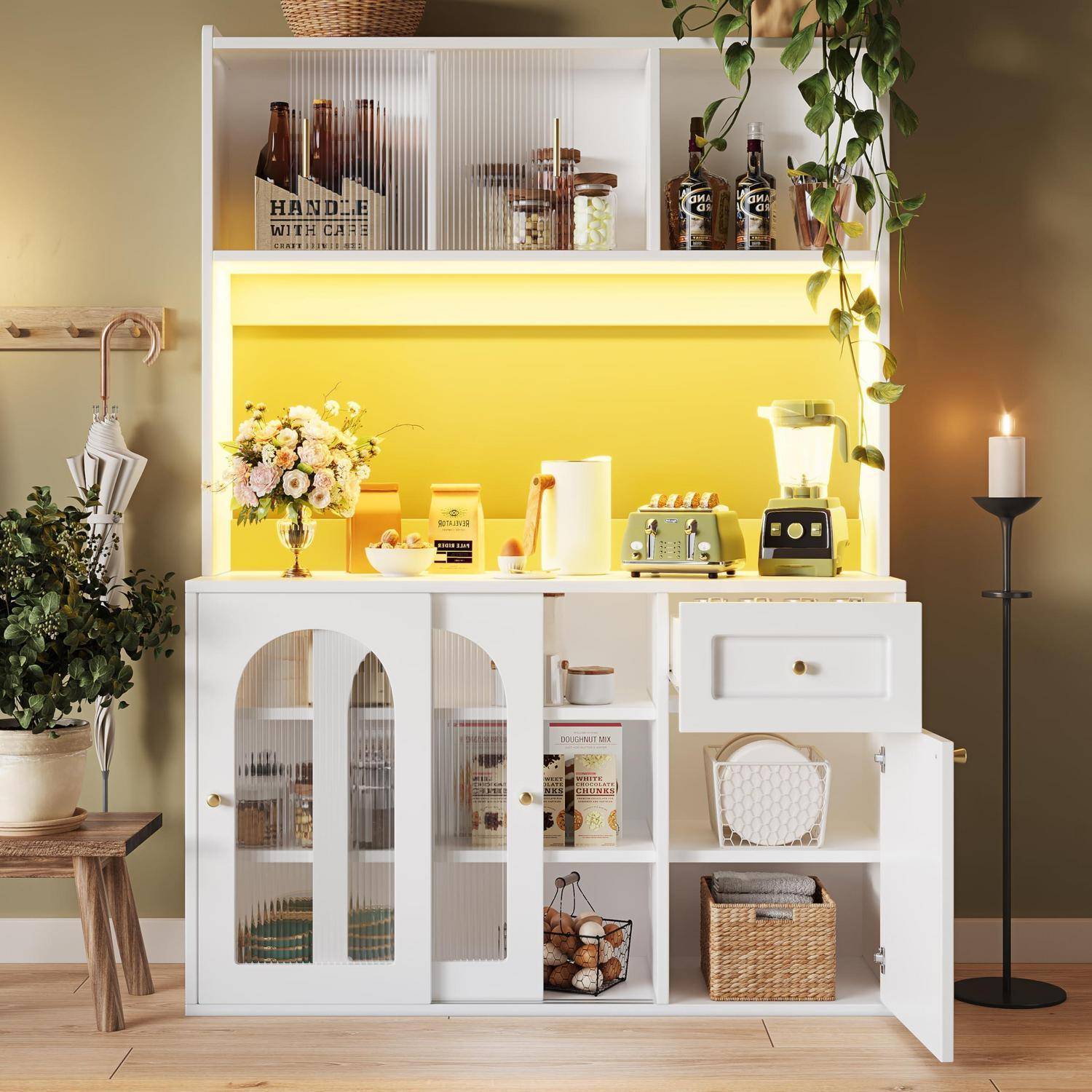 Weiße Küchenvitrине mit offenen und gläsernen Frontregalen, innen gelb beleuchtet, mit Kaffeeutensilien, Blumen, Toaster, Mixer, Körben und Dekoration.