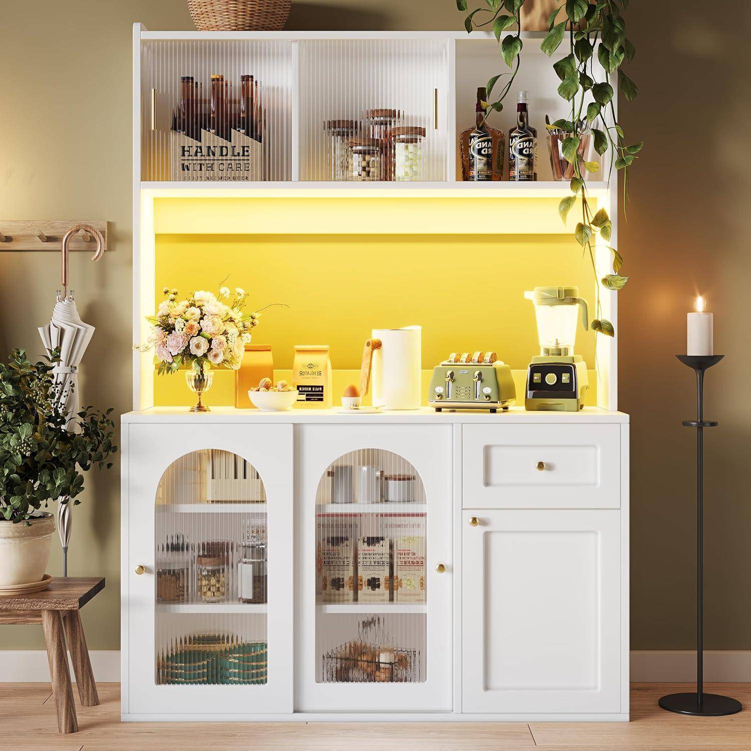 Weiße Küchenkommode mit gelb beleuchteter Rückwand zeigt Blumen, Gläser, Mixer, Toaster und Behälter; dekorative Heimatausstellung.