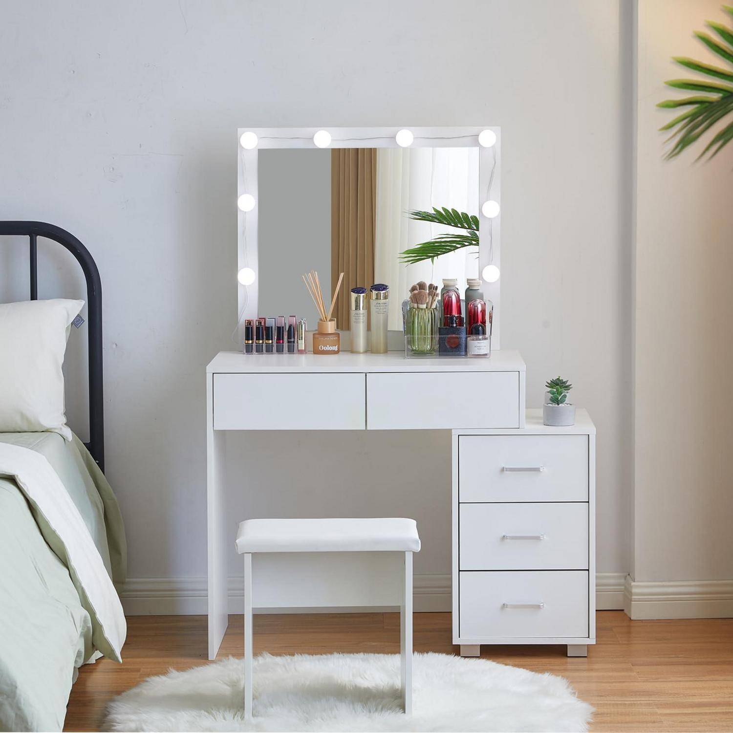 Weißer Frisiertisch mit beleuchtetem Spiegel, Makeup und Flaschen oben drauf, passender Hocker, neben einem Bett in einem hellen Schlafzimmer.