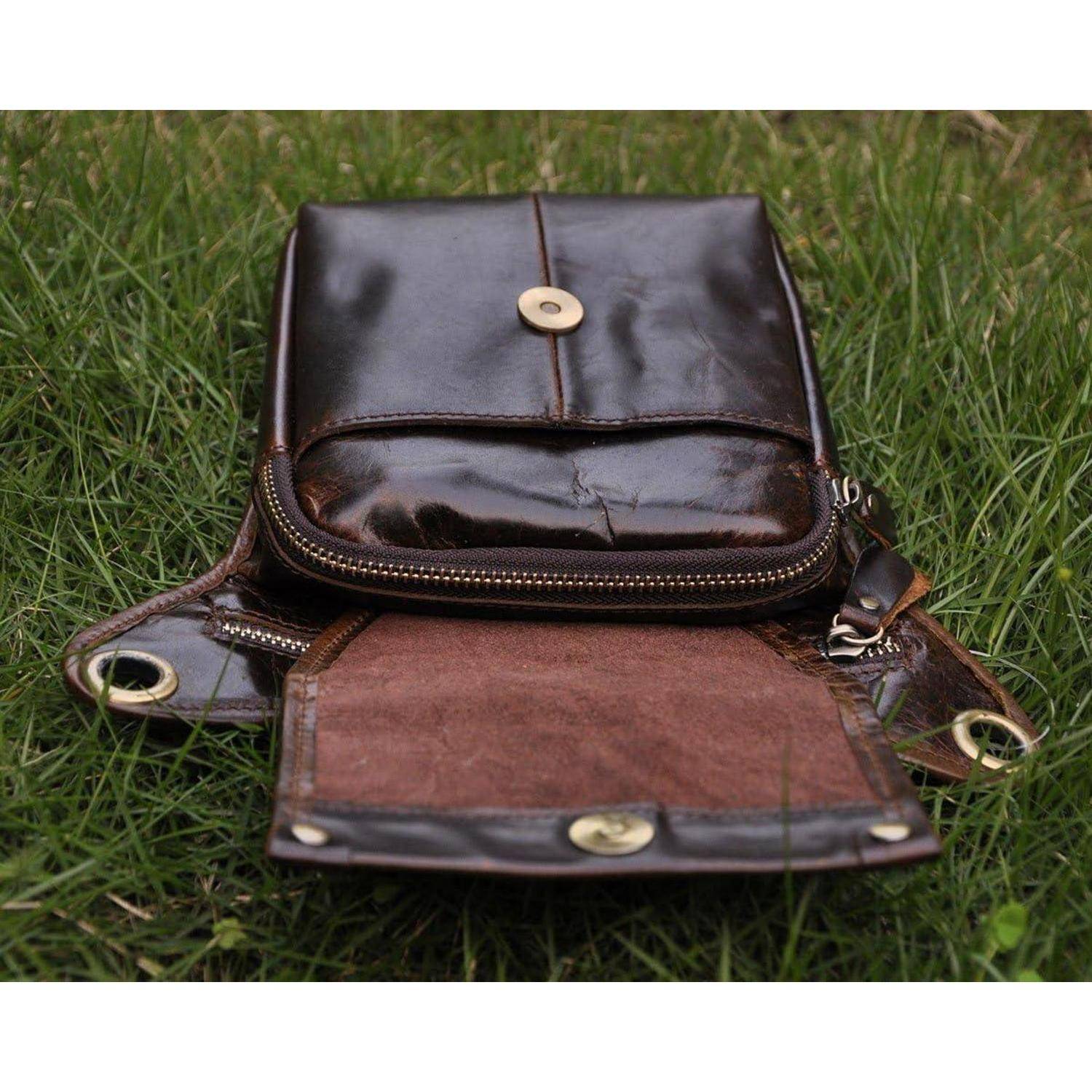 Braune Ledertasche oder kleiner Rucksack, offen auf Gras liegend, mit einer Frontlasche mit Reißverschluss und Messingknopfverschlüssen.