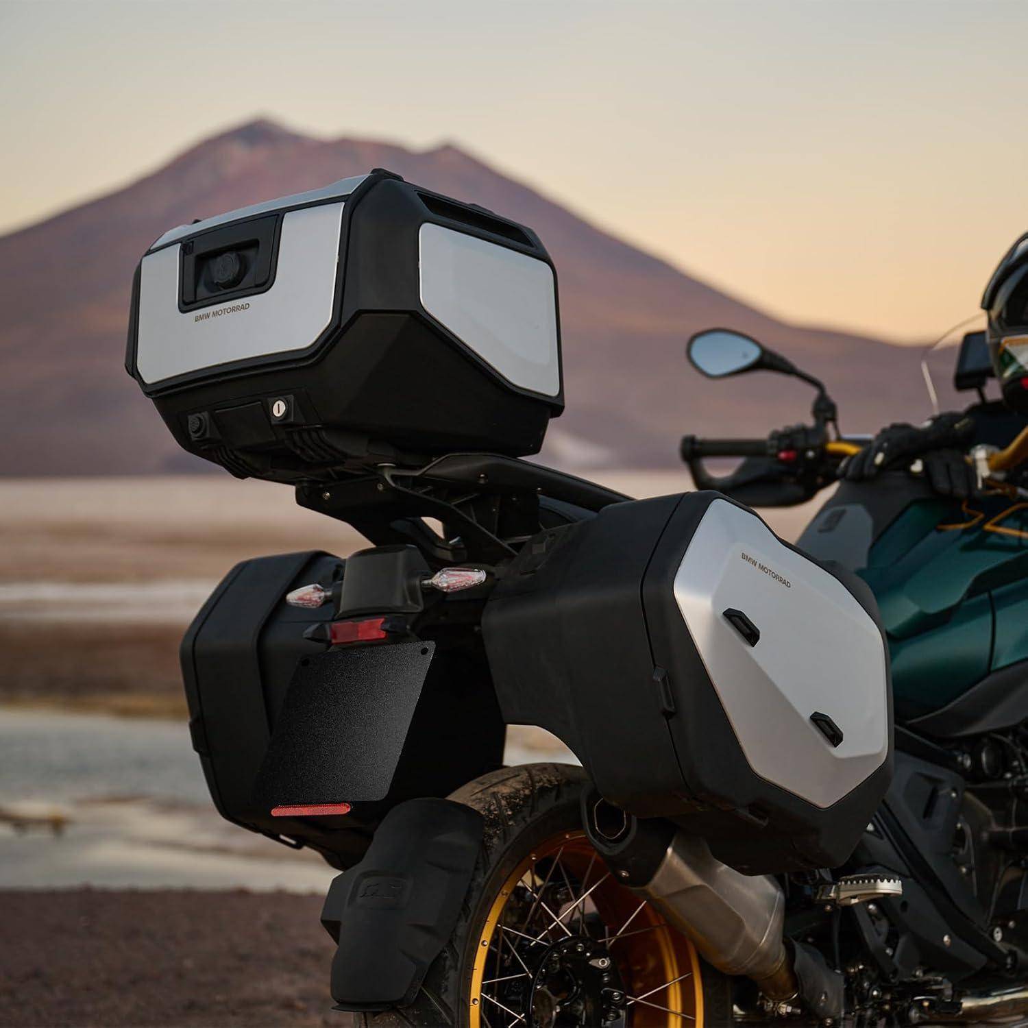 Tourenmotorrad mit hinteren hartschaligen Koffern, das am See bei Sonnenuntergang geparkt ist, mit Bergen im Hintergrund.