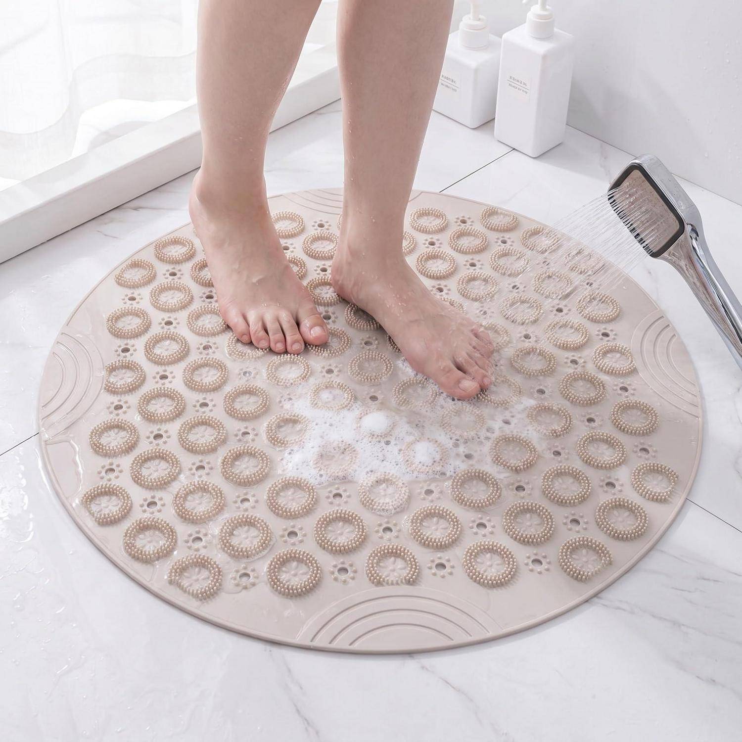 Eine Person steht auf einer runden beigefarbenen Badematte mit erhabenen Massageringen in einem weißen Badezimmer und spült ihre Füße mit einer tragbaren Handbrause ab.