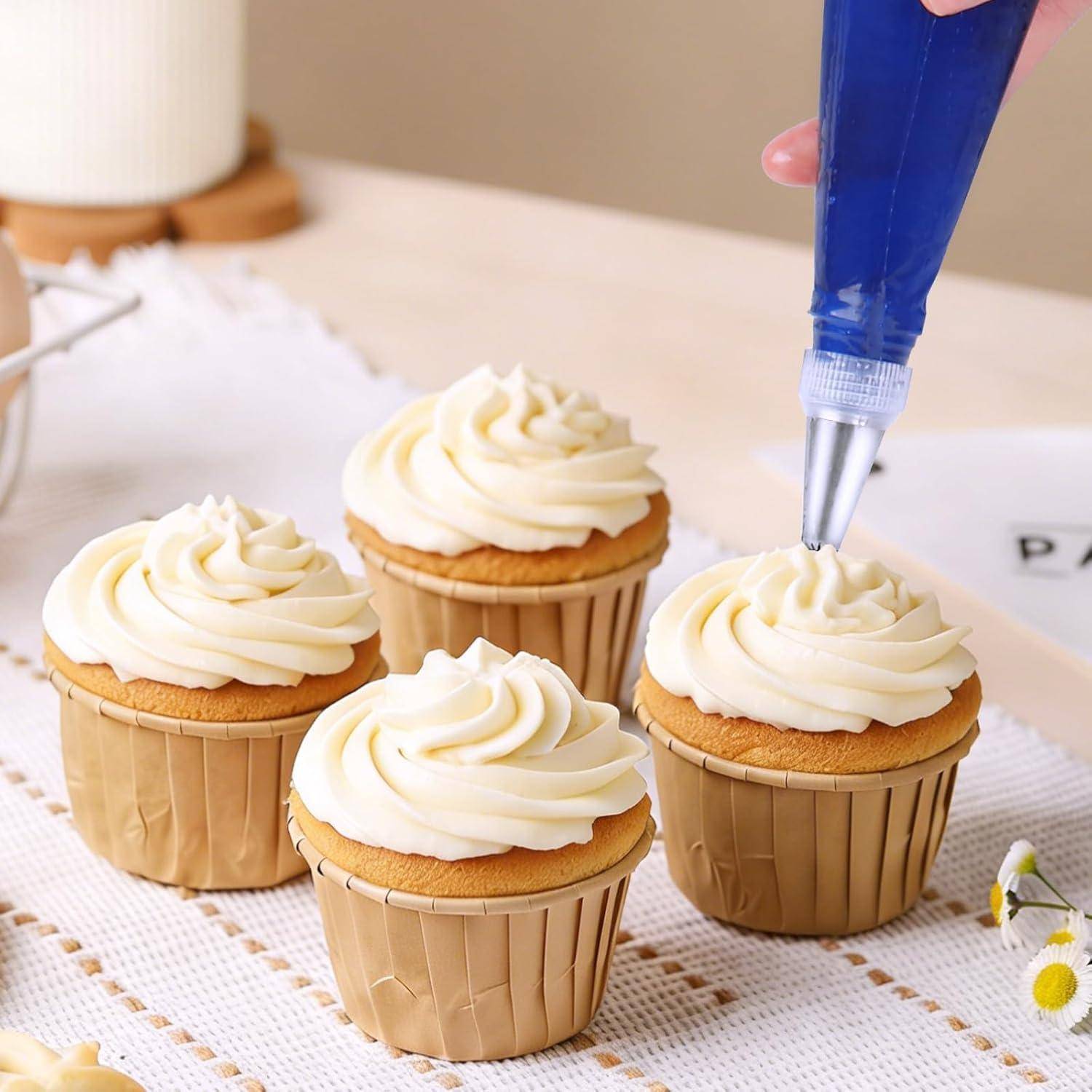Vier Cupcakes mit geschwungenem weißem Zuckerguss in beigen Förmchen, einer wird mit Zuckerguss aus einem blauen Spritzbeutel auf einer Küchentheke verziert.