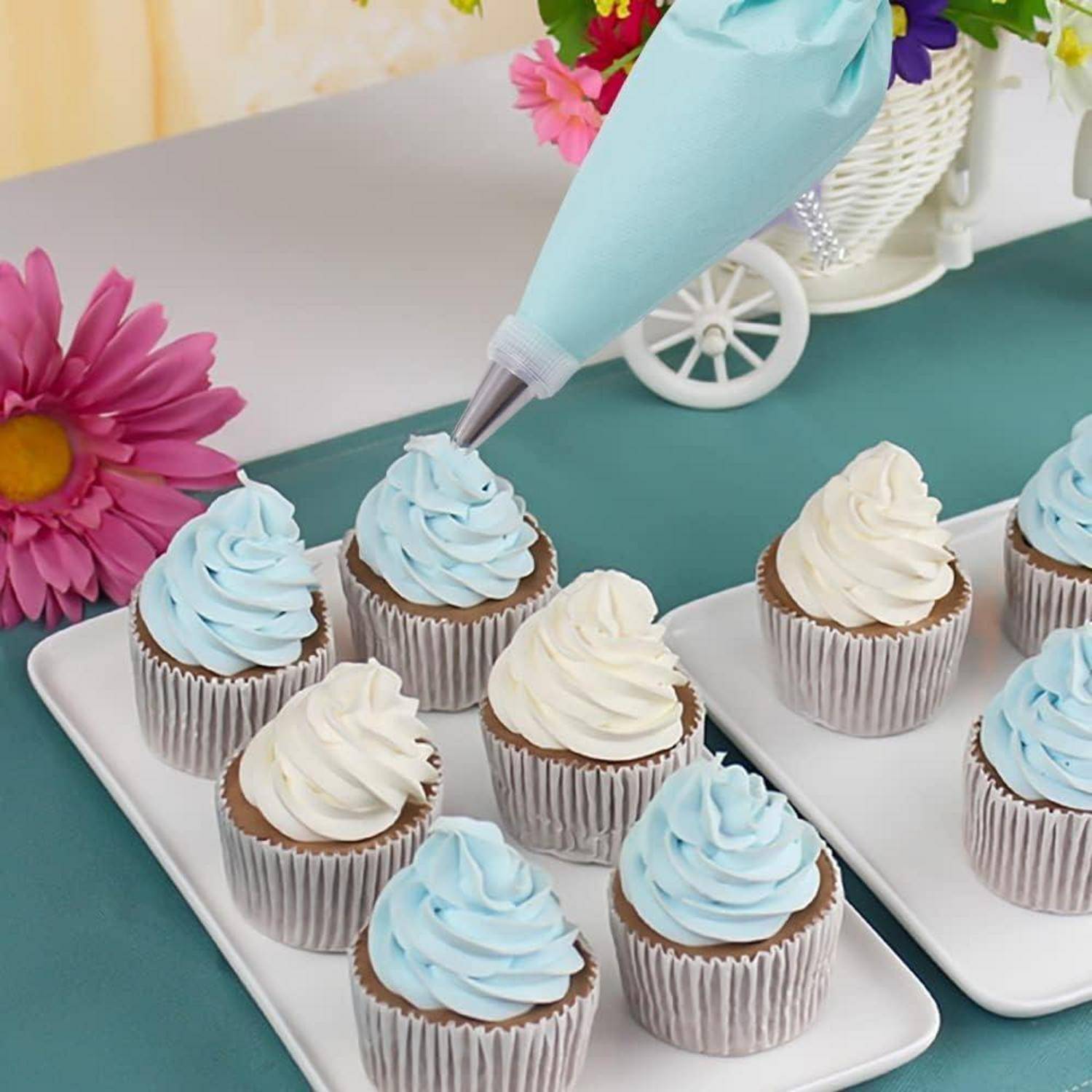 Hellblaue Glasur auf Cupcakes auf weißen Tabletts neben dekorierten Cupcakes und Blumen spritzen.
