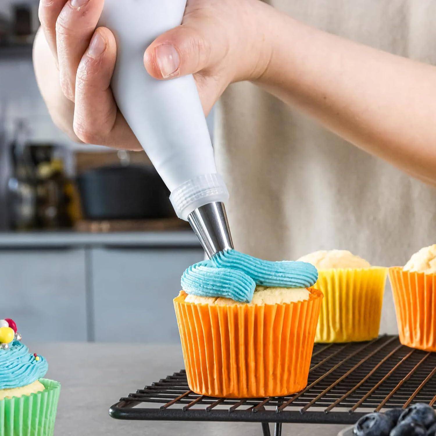 Eine Hand spritzt blaue Glasur mit einer Spritztüte auf einen Cupcake in einem orangefarbenen Papierförmchen, während weitere dekorierte Cupcakes auf einem Gitterrost abkühlen.