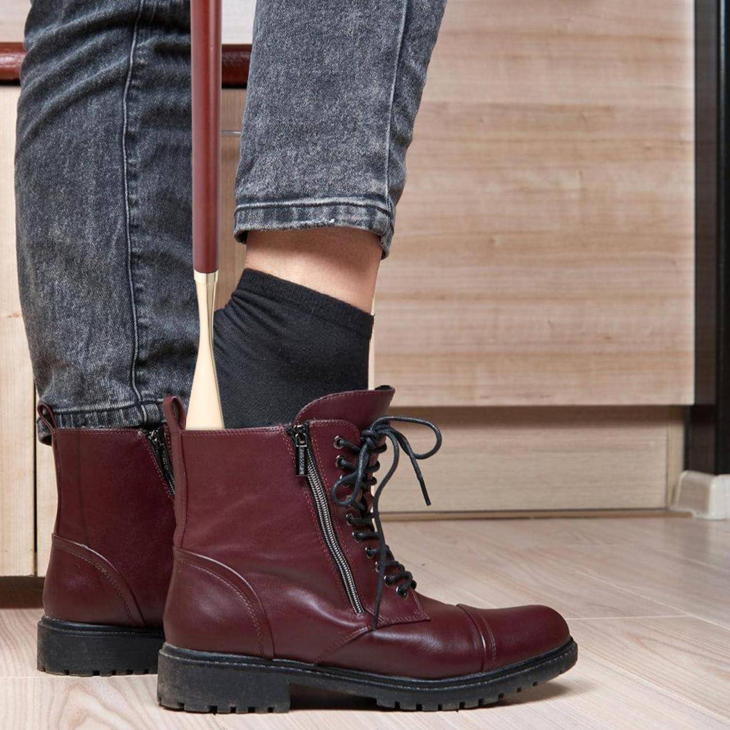 Person trägt weinrote Schnürstiefel im Knöchelbereich mit schwarzen Socken und hochgekrempelten Jeans und steht auf einem hellem Holzboden mit einem Besenstiel zwischen den Beinen.
