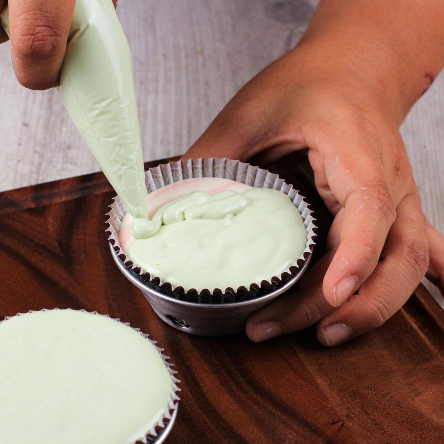 Hände, die hellgrüne Glasur mit einer Spritztüte in eine Cupcake-Form in einem Metallbecher auf einer Holzoberfläche spritzen, mit einem weiteren glasierten Cupcake in der Nähe.