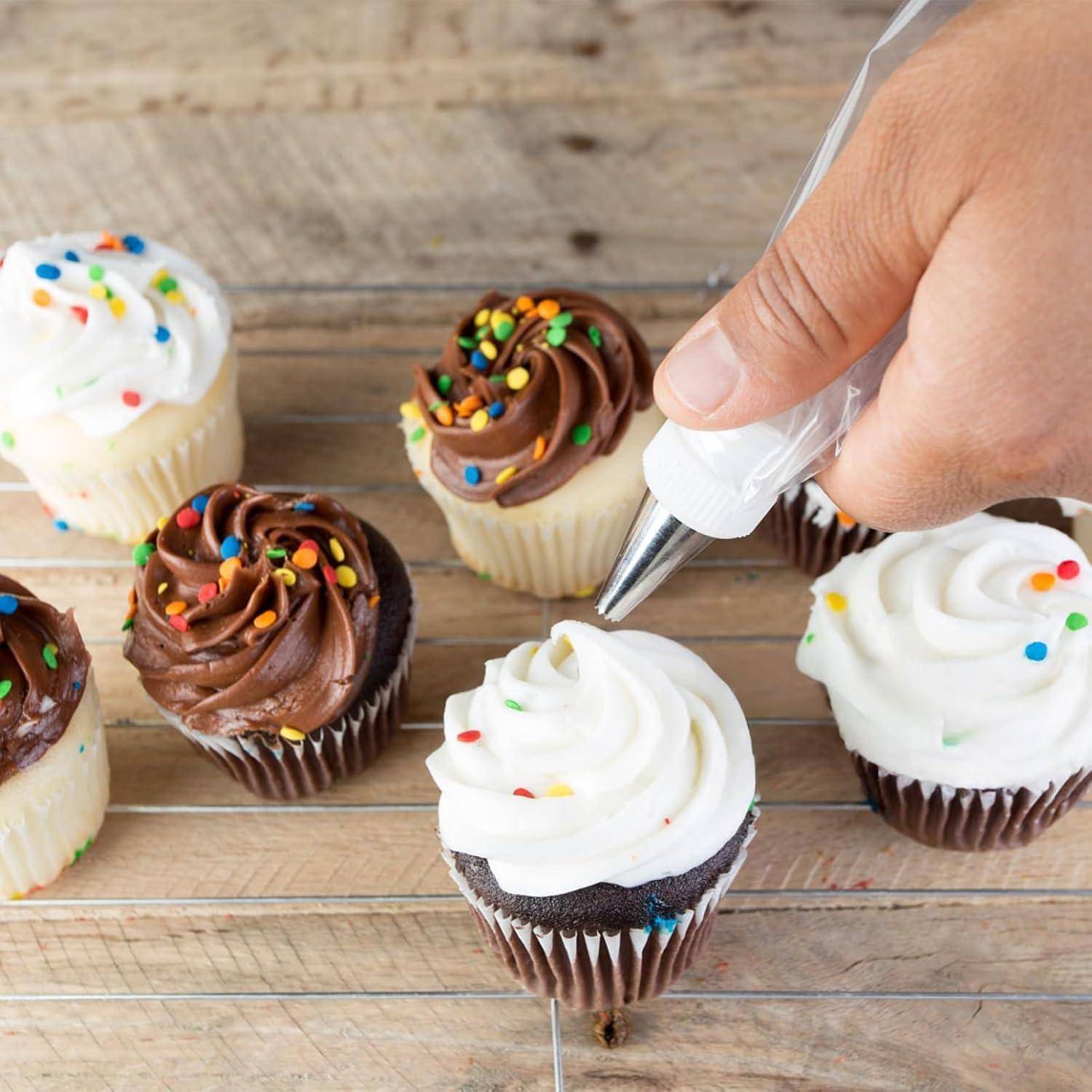 Mit der Hand weiße Glasur auf einen Schokoladen-Cupcake auf einem Holzbrett spritzen, mit mehreren dekorierten Cupcakes in der Nähe.