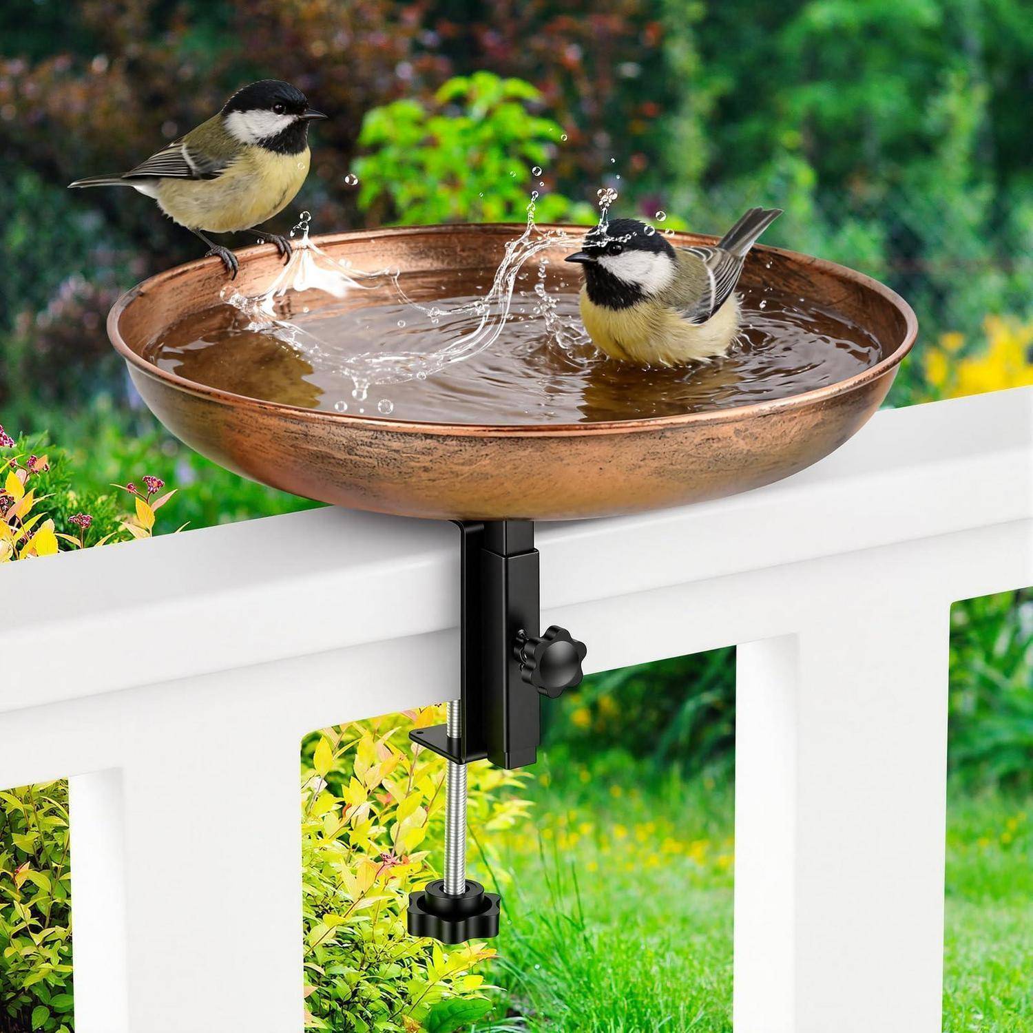 Zwei kleine Vögel sitzen auf einem Kupfervogelbad, das an einem weißen Zaun befestigt ist, während Wasser in der Schale spritzt und ein Garten hinter ihnen verschwommen ist.