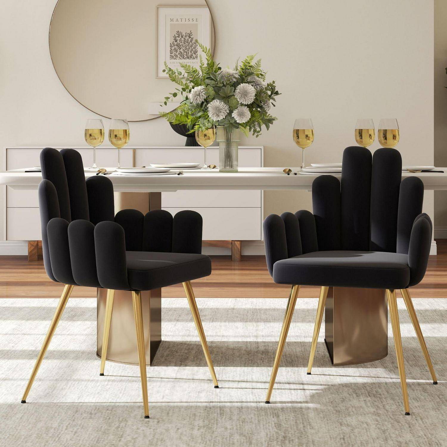 Modernes Esszimmer mit zwei schwarzen Samtstühlen mit goldfarbenen Beinen, die sich einer weißen Anrichte zuwenden, die mit Weingläsern und einer Blumenarrangement eingedeckt ist.