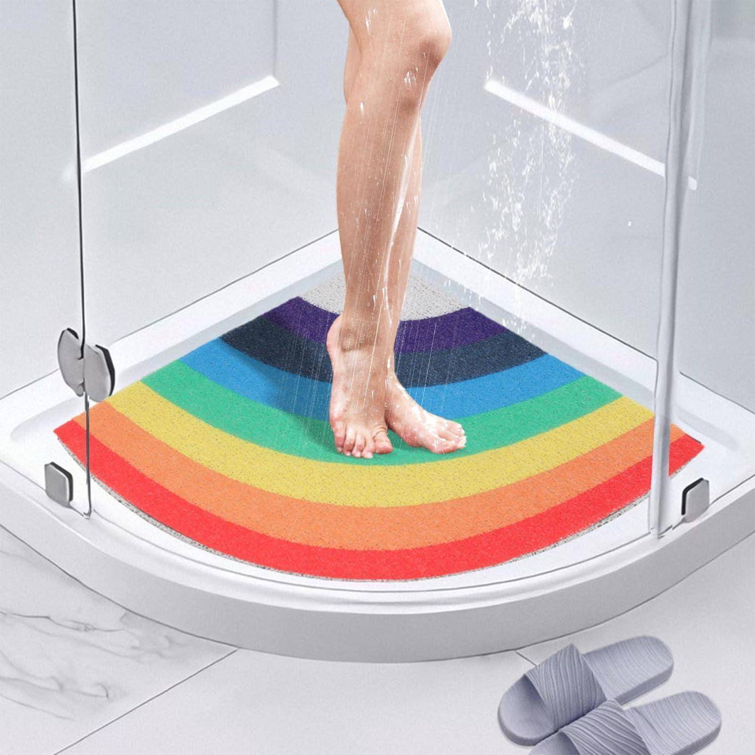 Person steht in einer gebogenen Glasdusche auf einer Regenbogen-Badematte, mit grauen Pantoffeln außerhalb auf einem weiß gefliesten Boden.