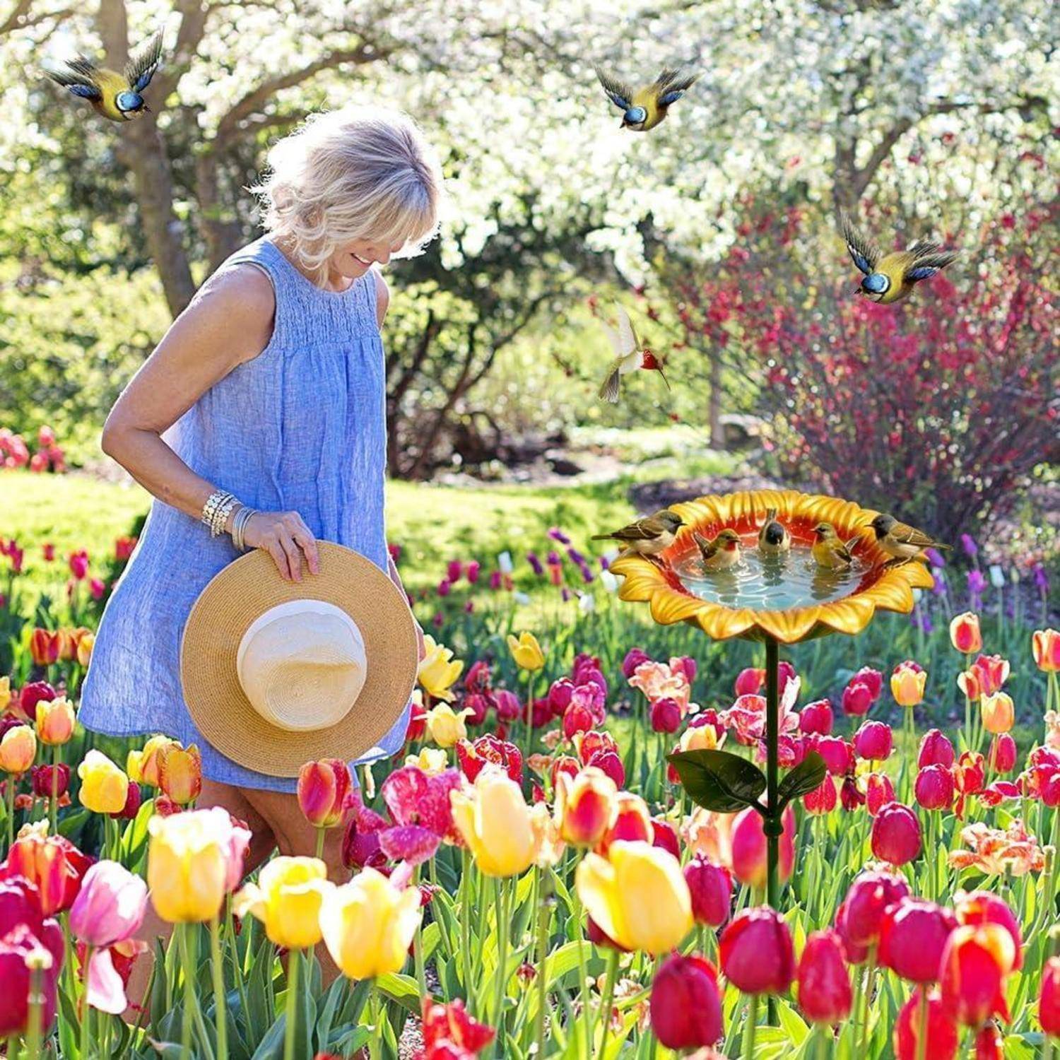 Eine Frau in einem blauen Kleid steht in einem farbenfrohen Tulpengarten, hält einen Strohhut und blickt nach unten; Vögel und ein Sonnenblumen-Vogelbad sind in der Nähe.