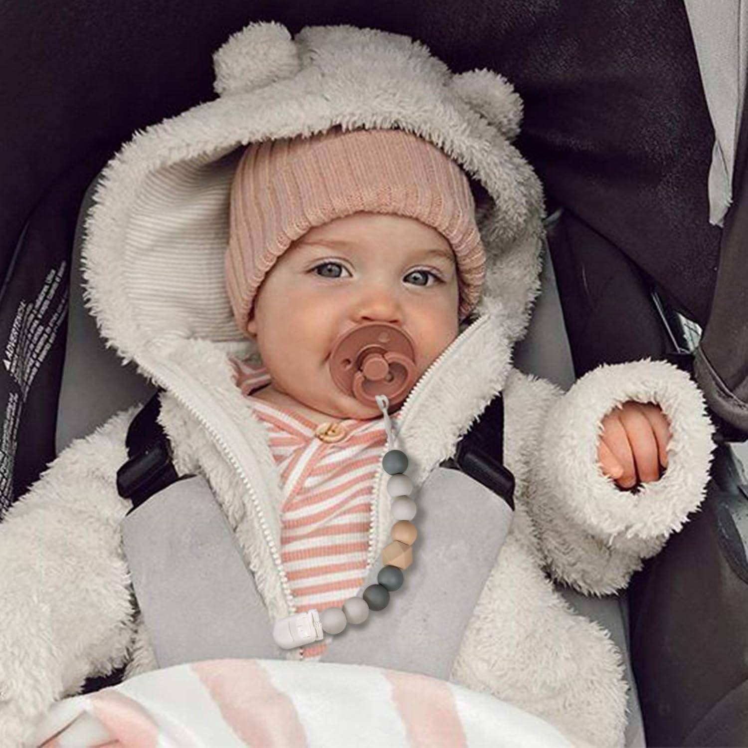 Baby in einem flauschigen Schneeanzug mit Kapuze und rosa Strickmütze sitzt in einem Kinderwagen mit einem Schnuller im Mund.