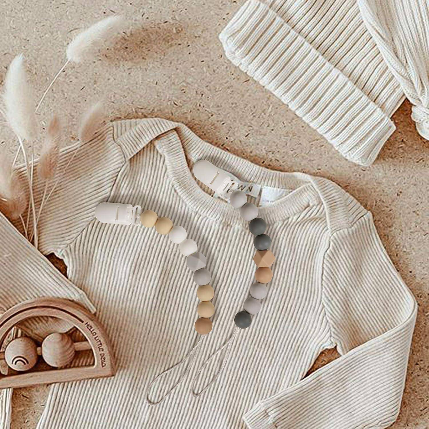 Cremefarbener gerippter Baby-Strampler mit abnehmbarem Silikon-Schnullerhalter, flach ausgelegt mit passendem Strickpflegenhut und Holzspielzeug für einen weichen, neutralen Kinderzimmerstil.