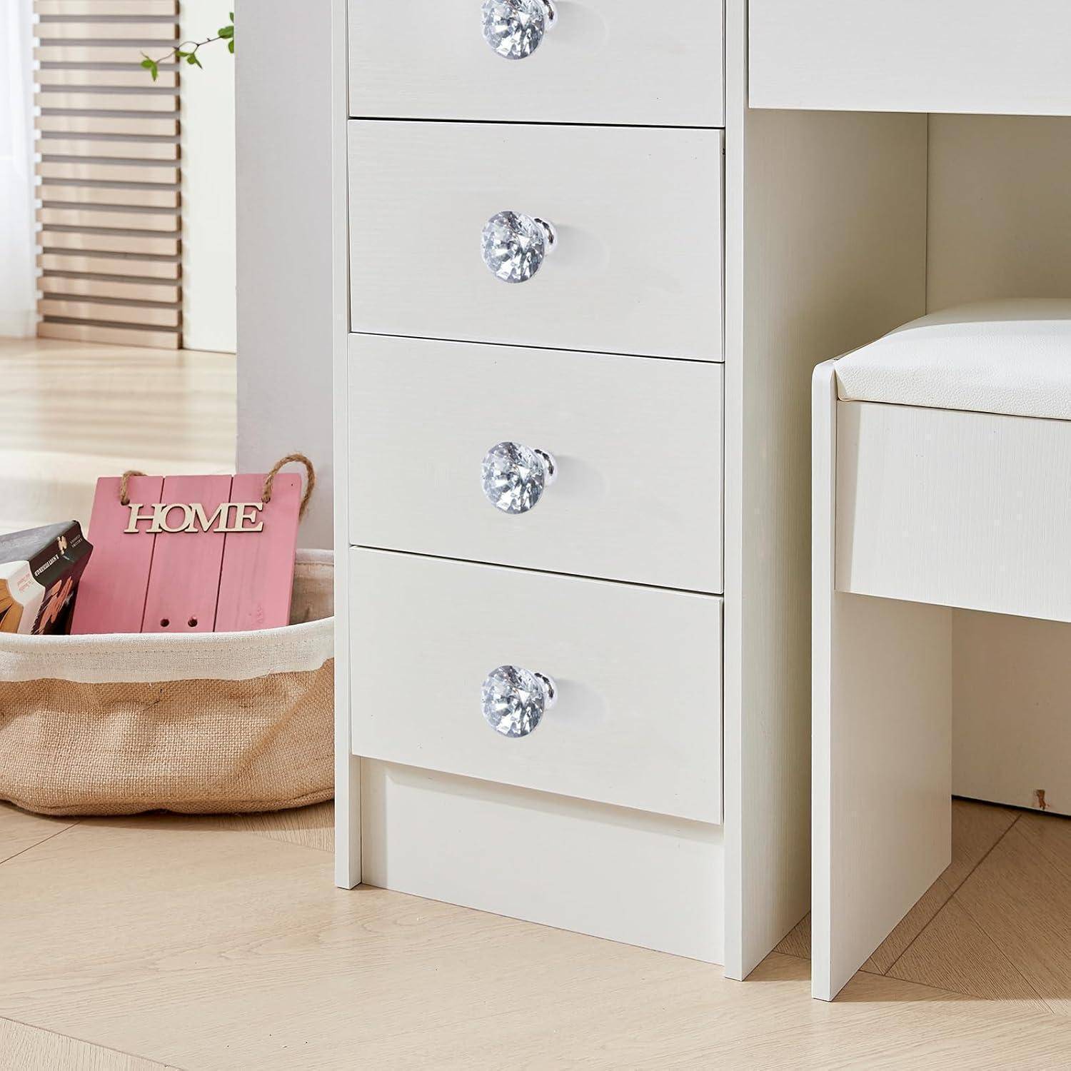 Weißer Schrank mit vier Schubladen und Kristallknöpfen neben einem passenden Schreibtisch; ein Korb und ein rosa Schild mit der Aufschrift „HOME" stehen auf dem Boden.