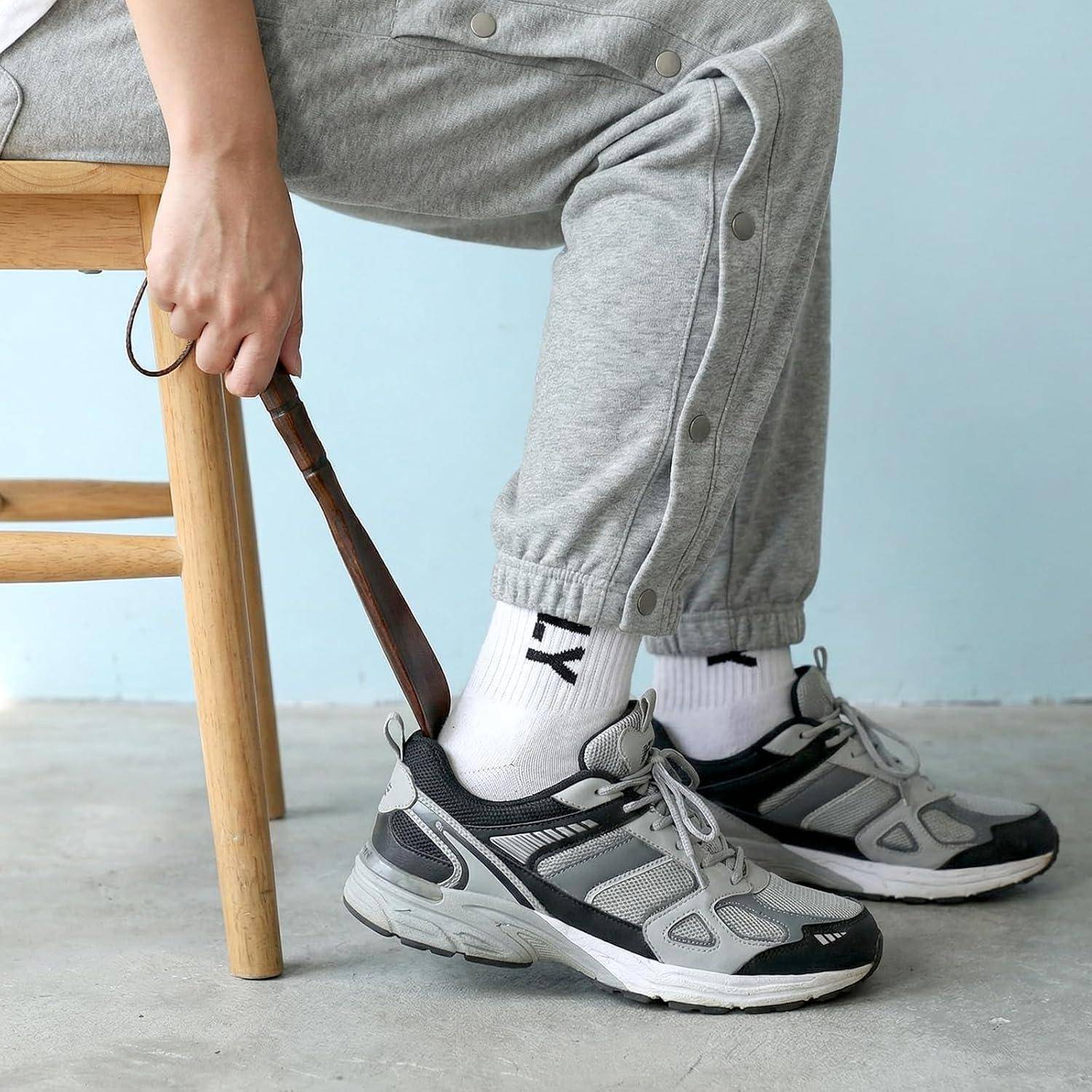 Eine Person sitzt auf einem hölzernen Stuhl und benutzt einen Schuhlöffel, um in grau-schwarze Sneaker zu schlüpfen, während sie eine graue Jogginghose und weiße Socken trägt.