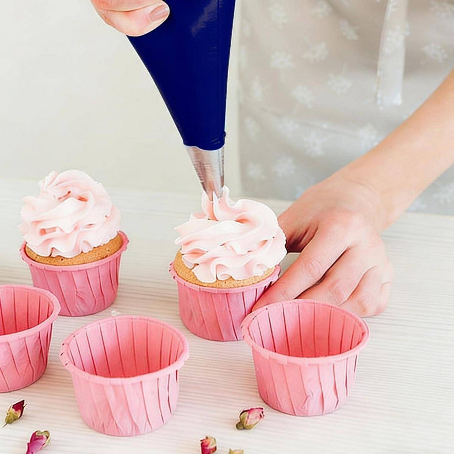 Person, die rosafarbenes Frosting auf Cupcakes in rosafarbenen Papierförmchen auf einem weißen Tisch spritzt.