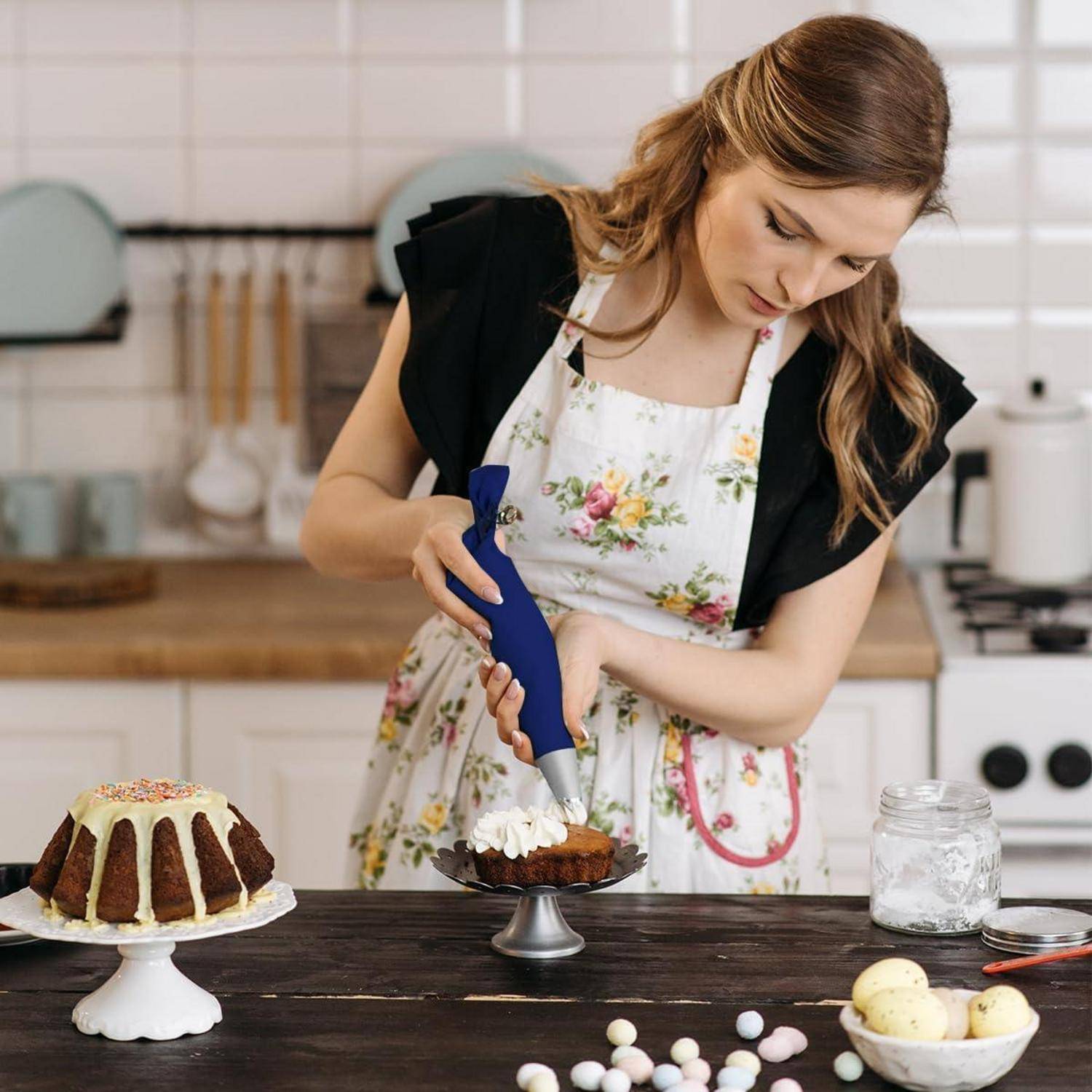Eine Frau dekoriert einen kleinen Schokoladenkuchen mit einem Spritzbeutel in einer Küche, mit einem größeren Gugelhupfkuchen, Zutaten und Küchenutensilien auf der Arbeitsplatte.