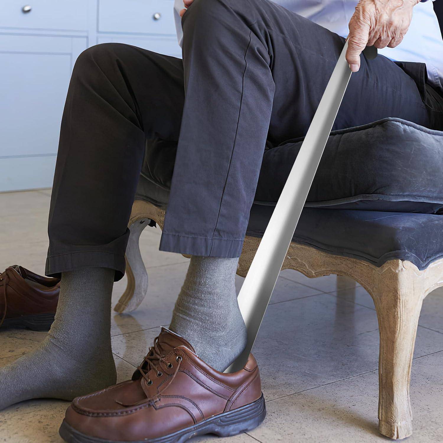 Eine ältere Person, die auf einem Stuhl sitzt, benutzt einen langen Schuhlöffel, um braune Loafer über grauen Socken anzuziehen.