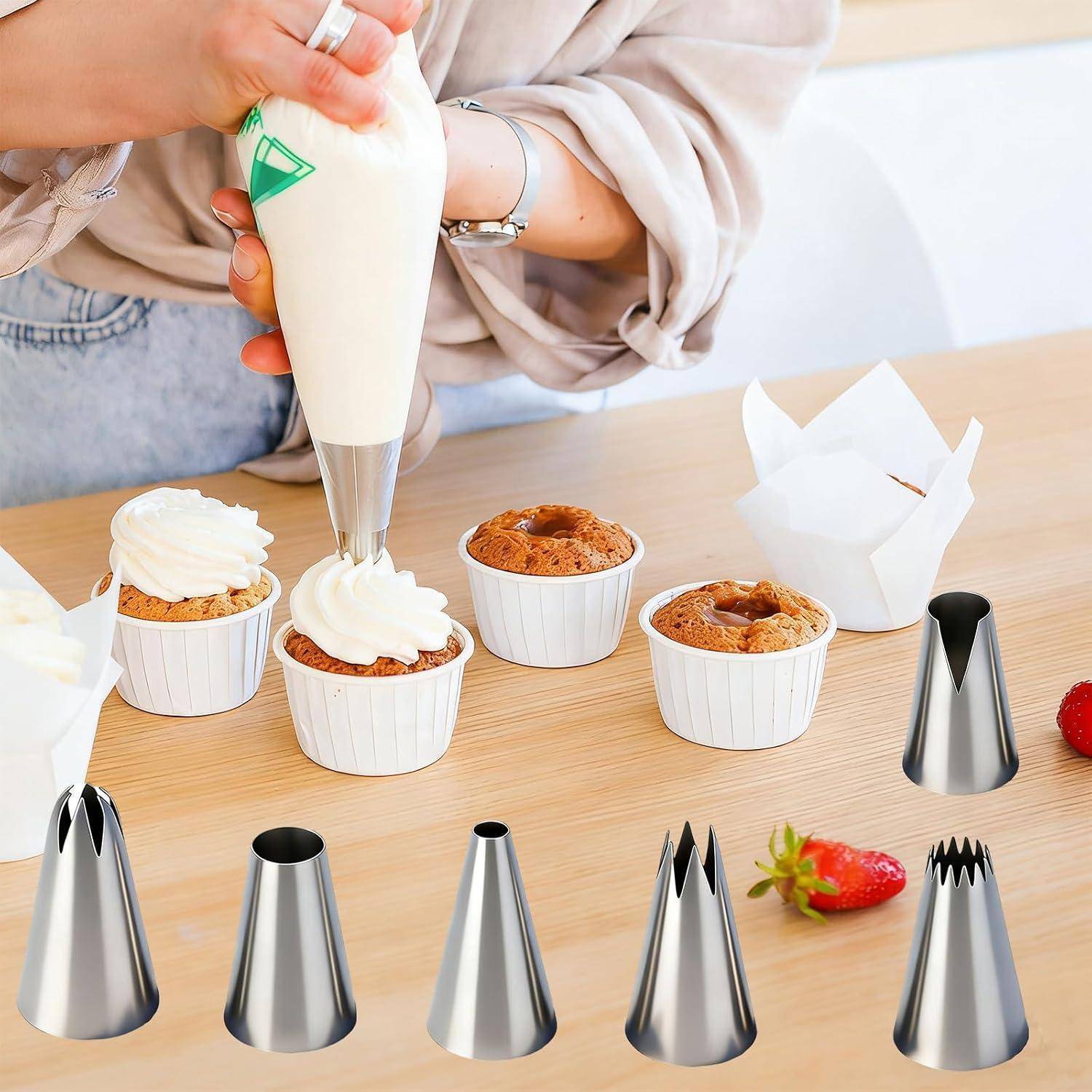 Person, die weiße Glasur mit einem Spritzbeutel auf Cupcakes in weißen Auflaufförmchen aufträgt, mit mehreren Metall-Spritztüllen und Erdbeeren auf einem Holztisch.