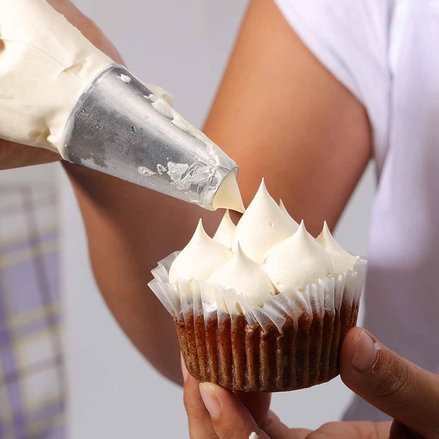 Ein Bäcker spritzt weiße Glasur mit einem Spritzbeutel aus Metall auf einen in der Hand gehaltenen Cupcake und formt damit spitz zulaufende Wirbel.