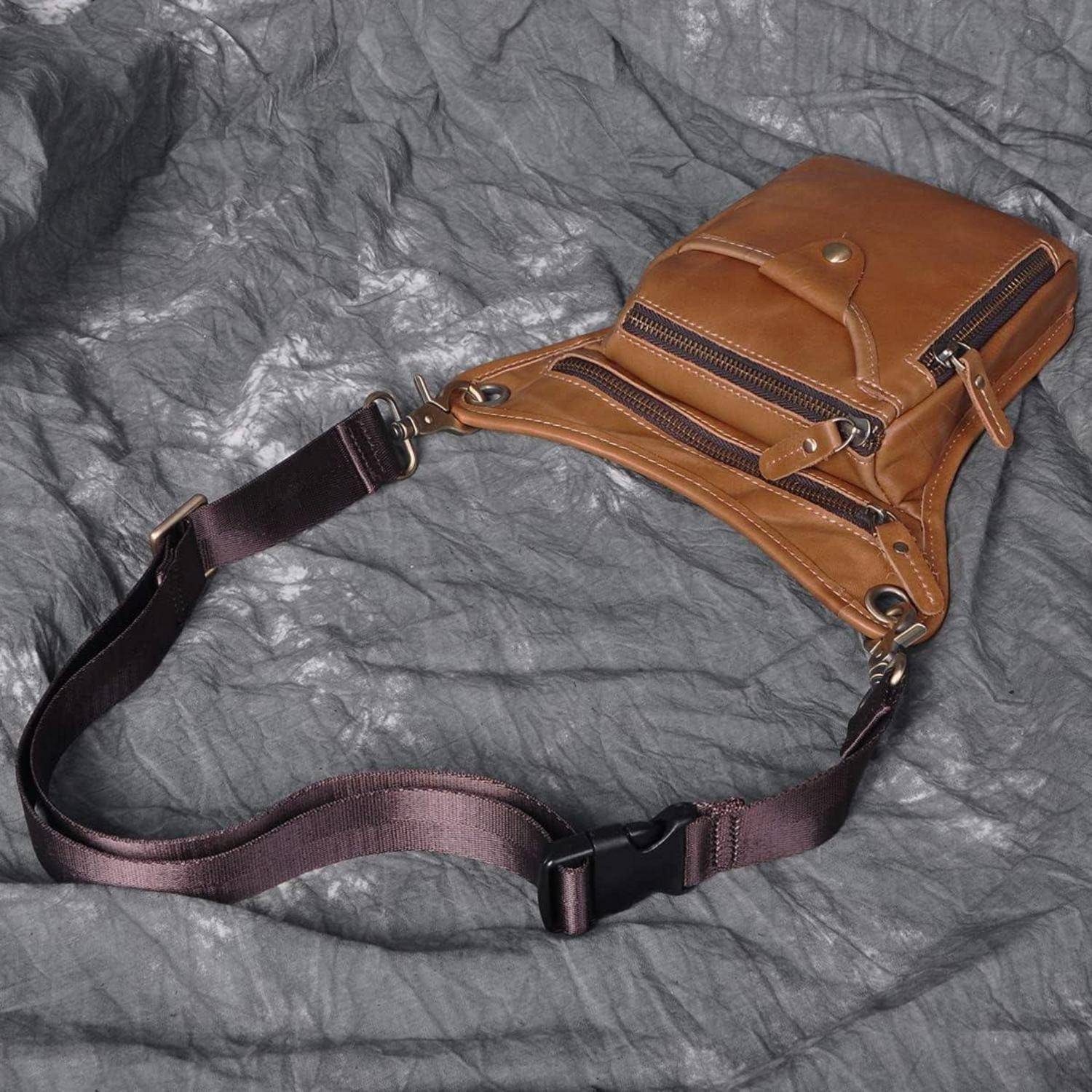 Braune Ledertasche zum Umhängen mit zwei vorderen Reißverschlüssen und verstellbarem dunklem Riemen auf grauem, zerknittertem Stoffhintergrund.