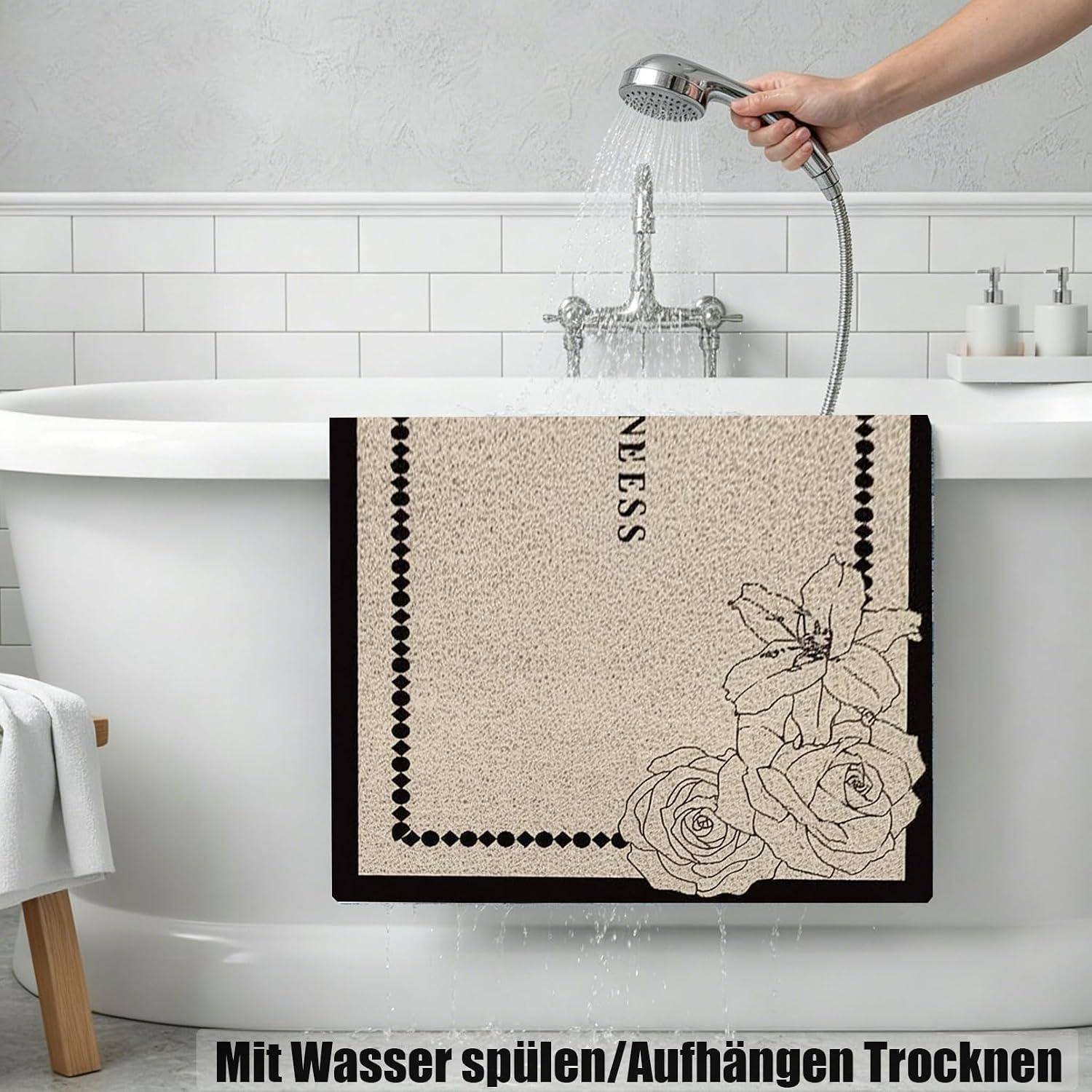 Hand, die einen Duschkopf hält und eine beigefarbene Badematte spült, die über einer Badewanne drapiert ist, mit deutschem Text „Mit Wasser spülen/Aufhängen Trocknen