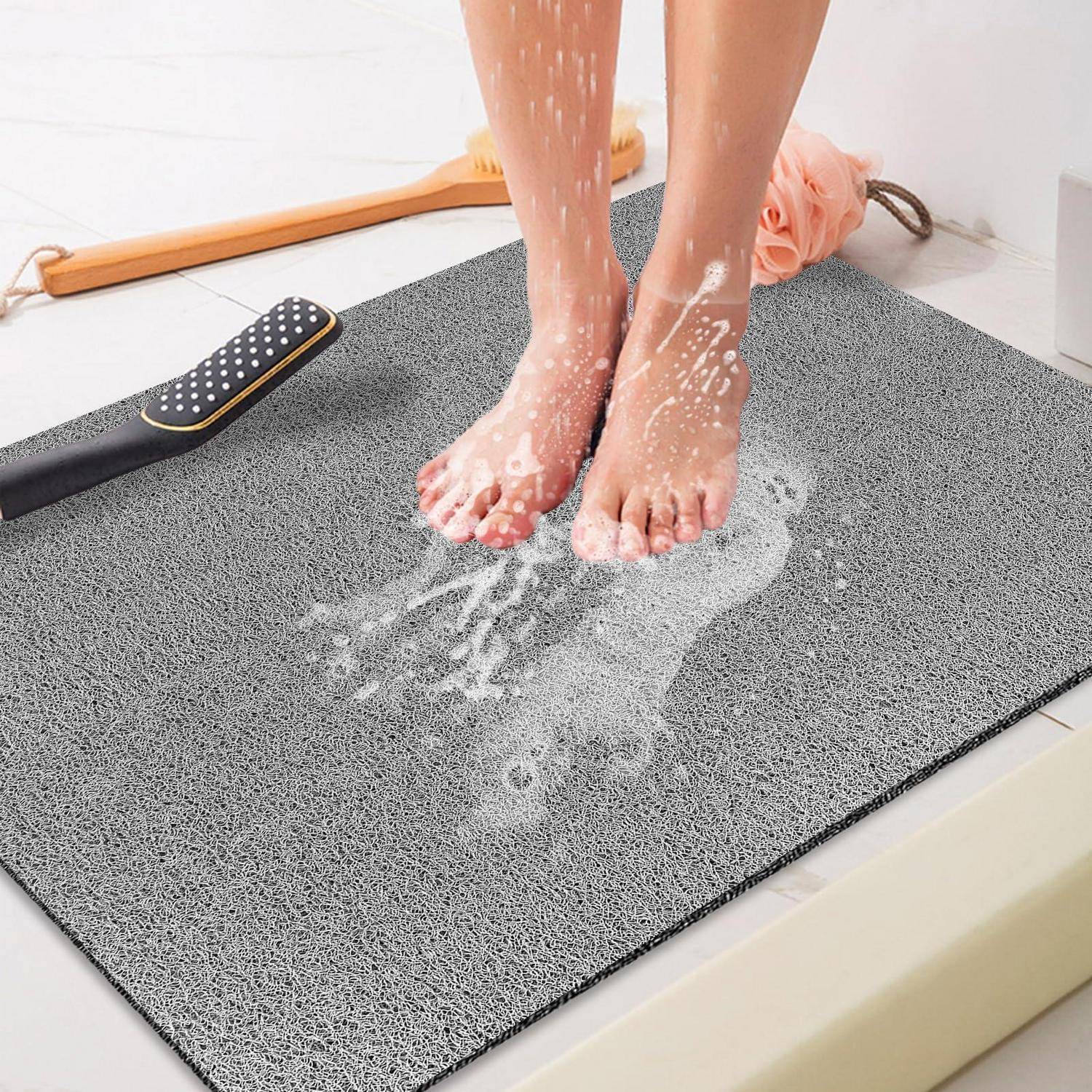 Nackte Füße, die auf einer grauen saugfähigen Badematte stehen, während Wasser spritzt und abfließt, mit einer Bürste und einem Handtuch im Hintergrund.