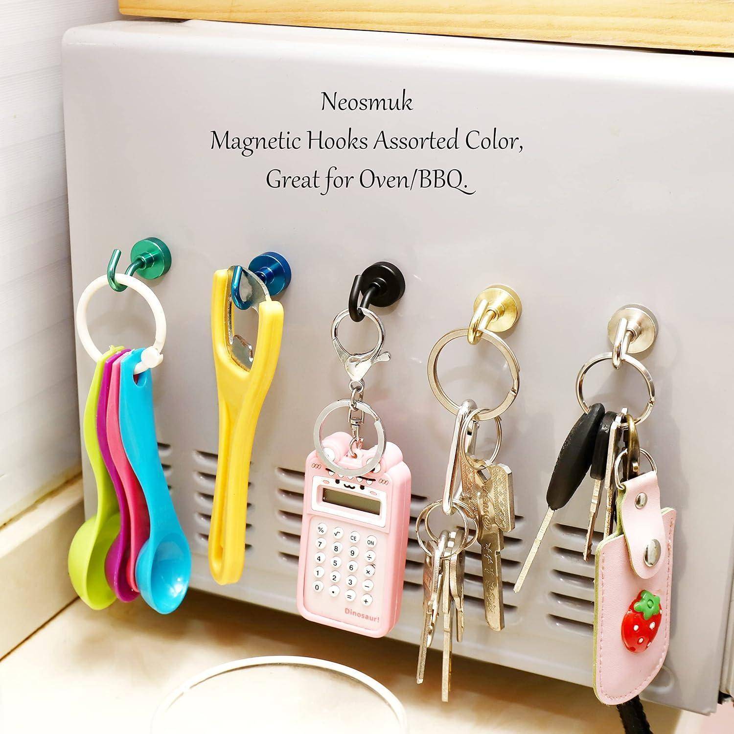 Produktfoto von Neosmuk-Magnethaken in verschiedenen Farben, die Küchenutensilien, Schlüssel und kleine Accessoires an einer Metalloberfläche halten.