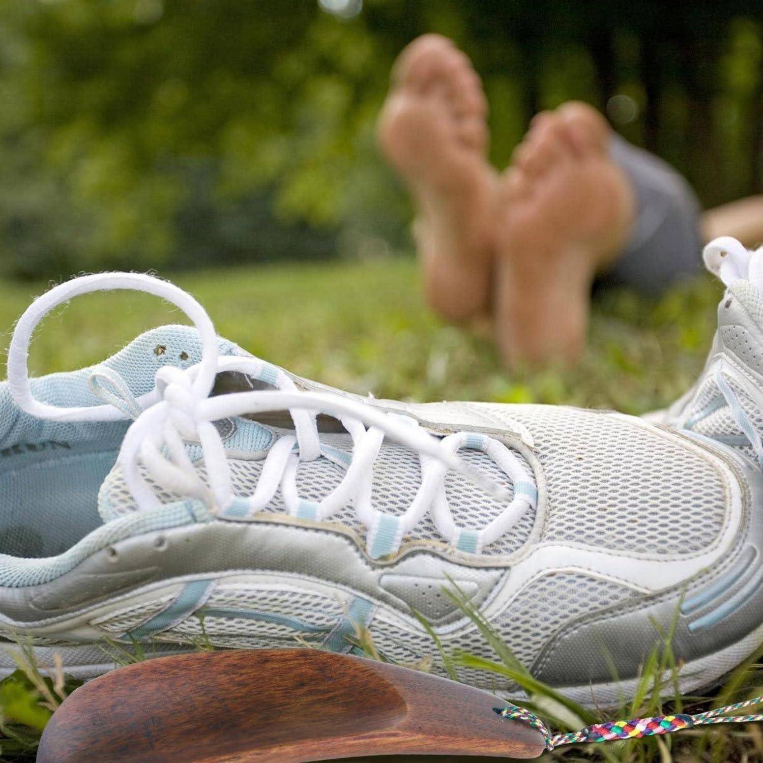 Nahaufnahme von weißen Laufschuhen auf Gras, mit einer Person, die barfuß im unscharfen Hintergrund sitzt.
