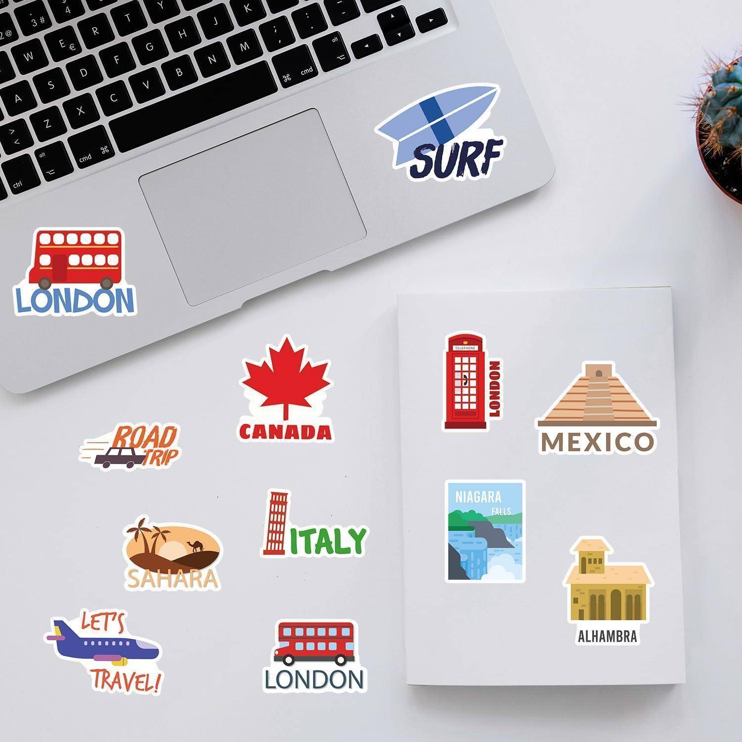 Reiseaufkleber auf einem Schreibtisch neben einem Laptop und Notizbuch, die Reiseziele und Symbole mit den Beschriftungen London, Surf, Road Trip, Kanada, Sahara, Italien, Mexiko, Niagarafälle und Alhambra zeigen.