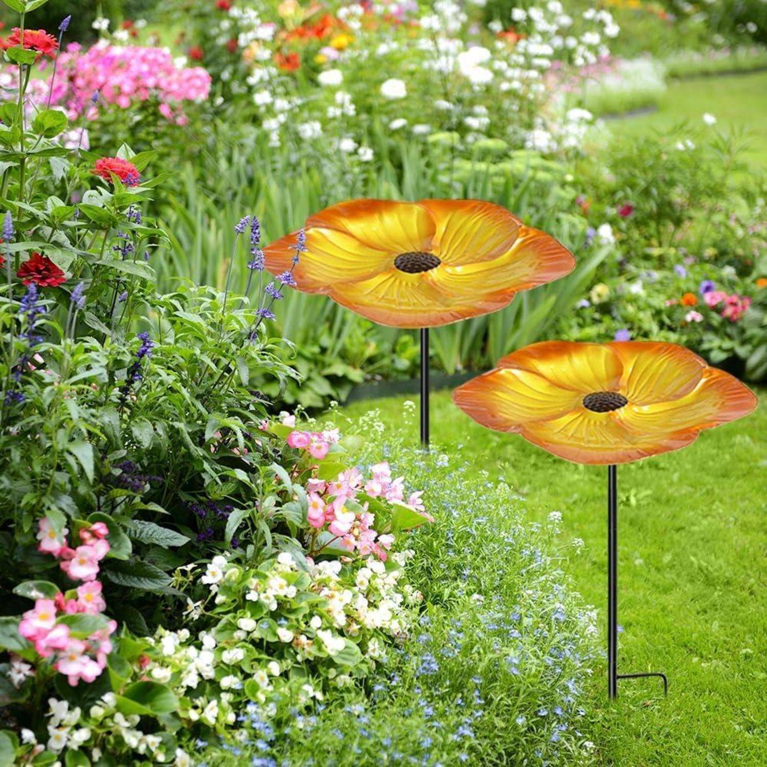 Garten mit zwei orangefarbenen blütenförmigen Metallskulpturen auf Stäben zwischen farbenfrohen blühenden Pflanzen und grünem Rasen.